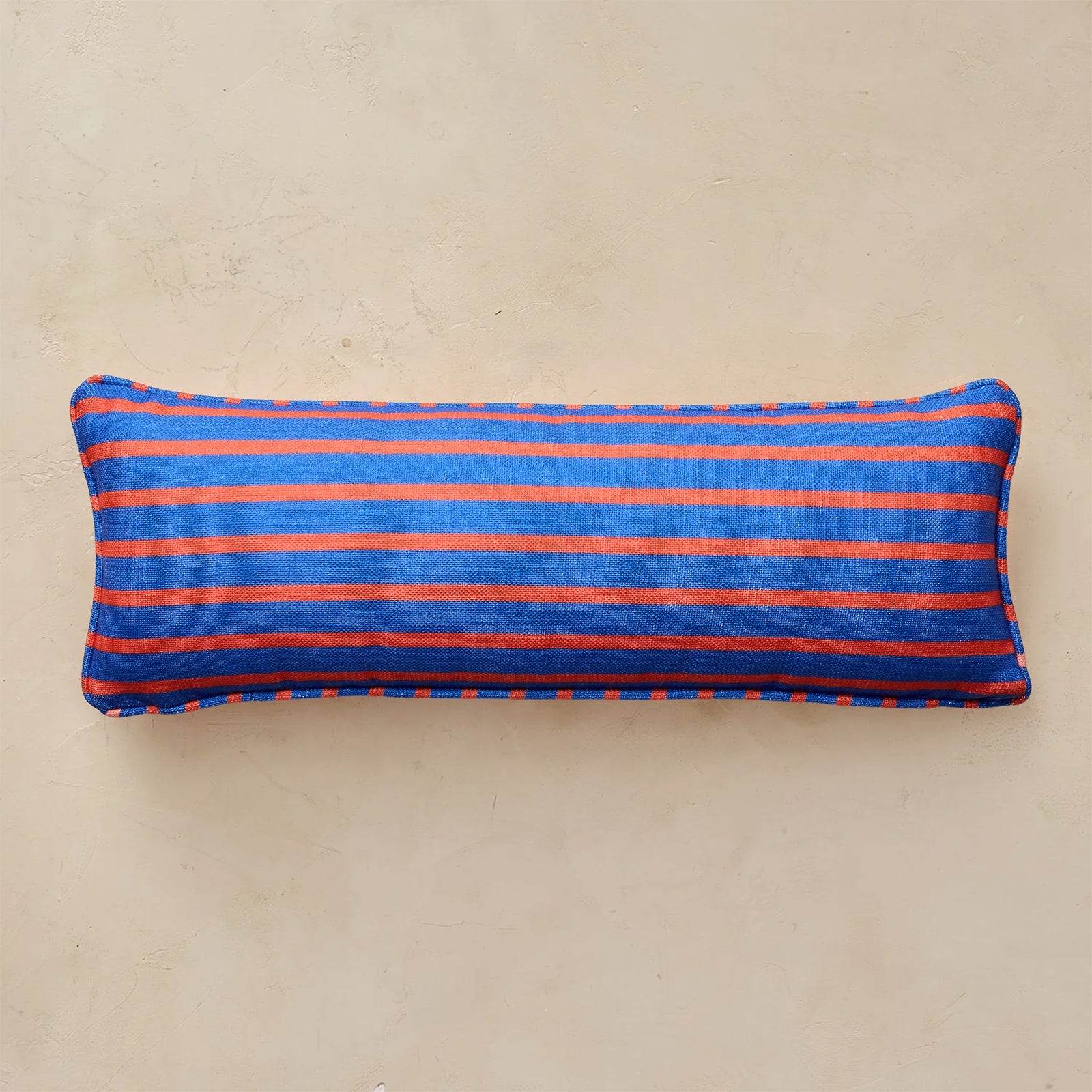 A rectangular lumbar pillow with horizontal stripes in royal blue and orange, resting on a textured beige surface.