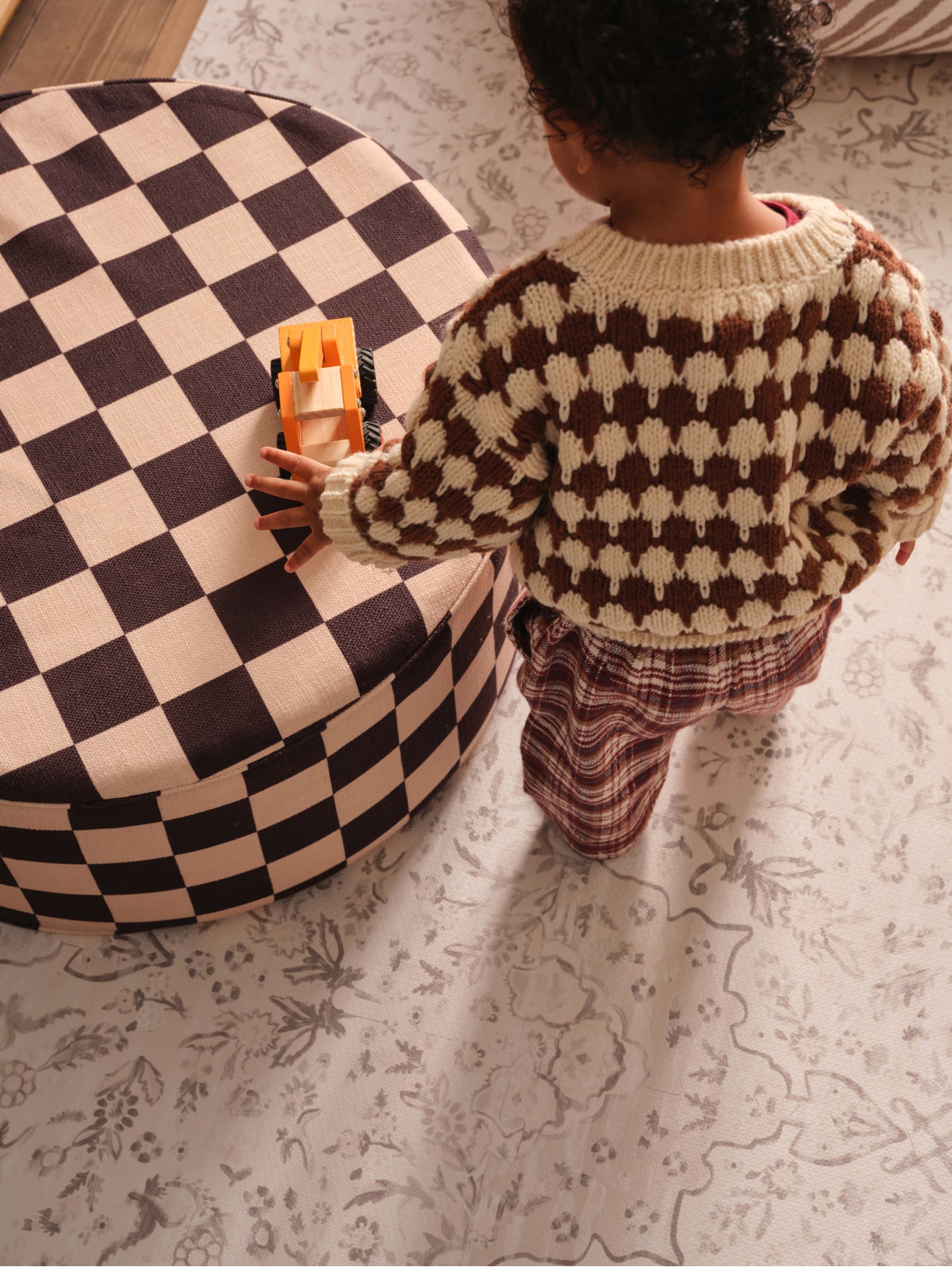 Child in a checkered sweater playing with a toy truck on a checkered ottoman on a play mat.