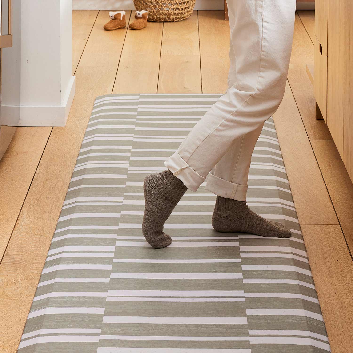sutton stripe basil olive green and white inverted stripe standing mat shown in kitchen with womans legs in white jeans and gray socks standing on the mat