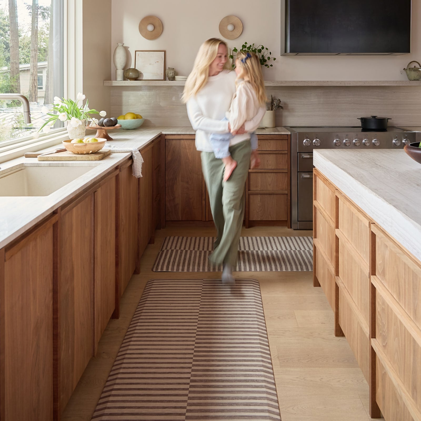 Woman and child in a modern kitchen with wooden cabinets and a striped kitchen mat.