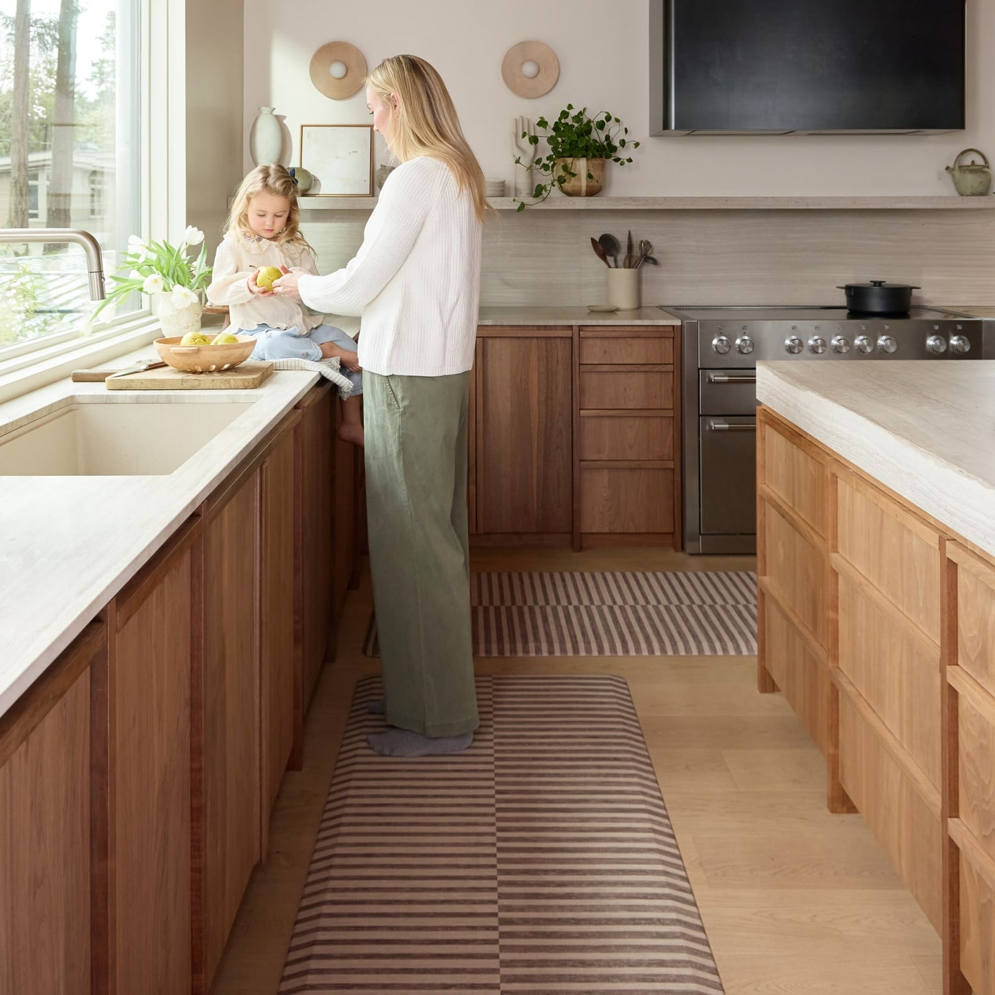 Woman and child in a modern kitchen with wooden cabinets and a striped kitchen mat.