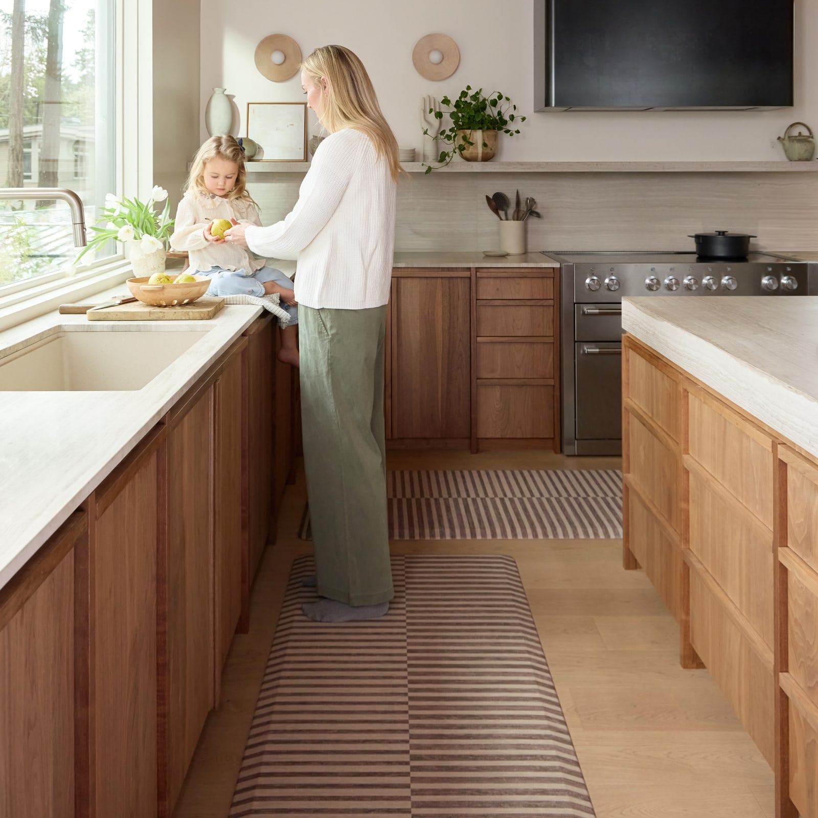 Woman and child in a modern kitchen with wooden cabinets and a striped kitchen mat.