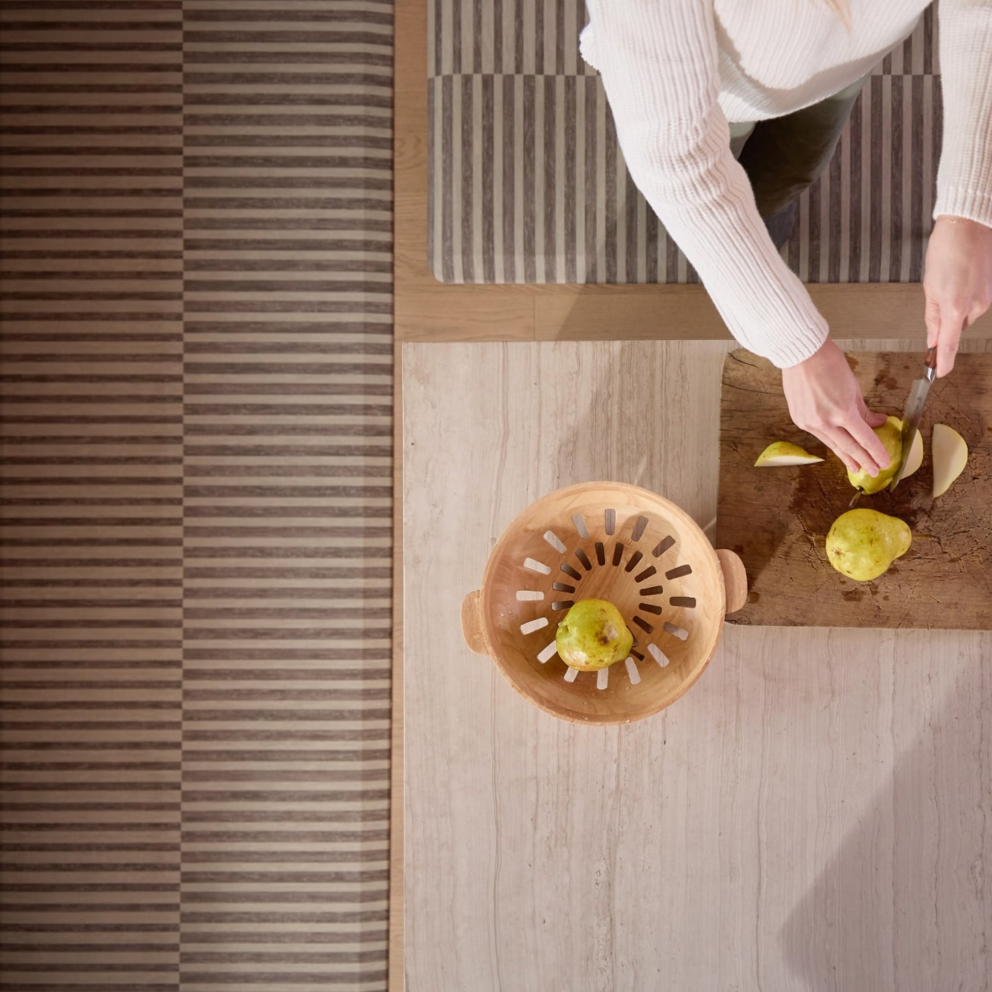 Person cutting pears on a wooden cutting board standing on a striped kitchen mat with a wooden colander nearby.