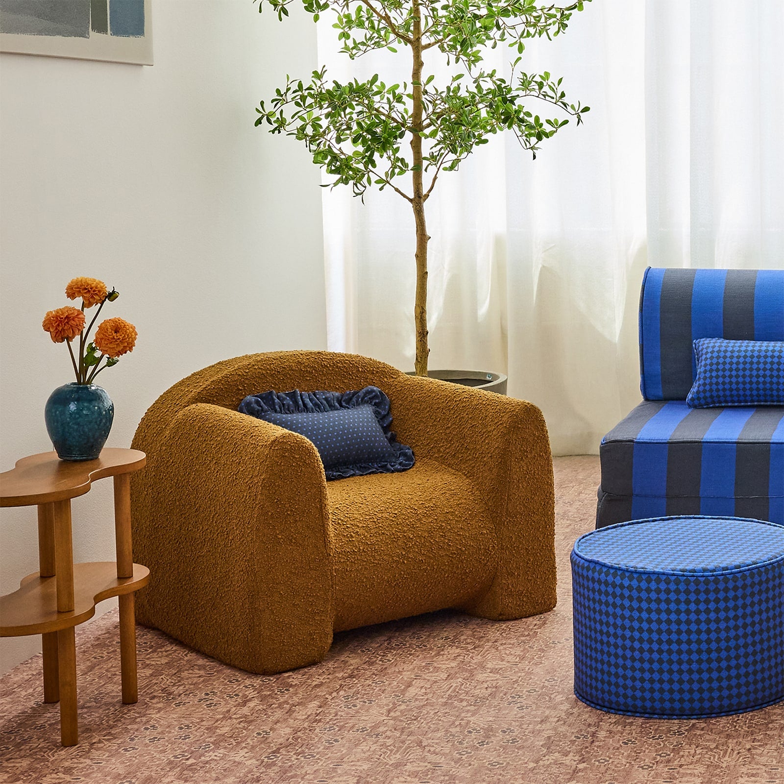 A cozy living room scene featuring a textured rust brown armchair with a blue pillow, a blue-and-navy striped play couch, a blue patterned ottoman, a small wooden side table with orange flowers, and a potted tree in the background.