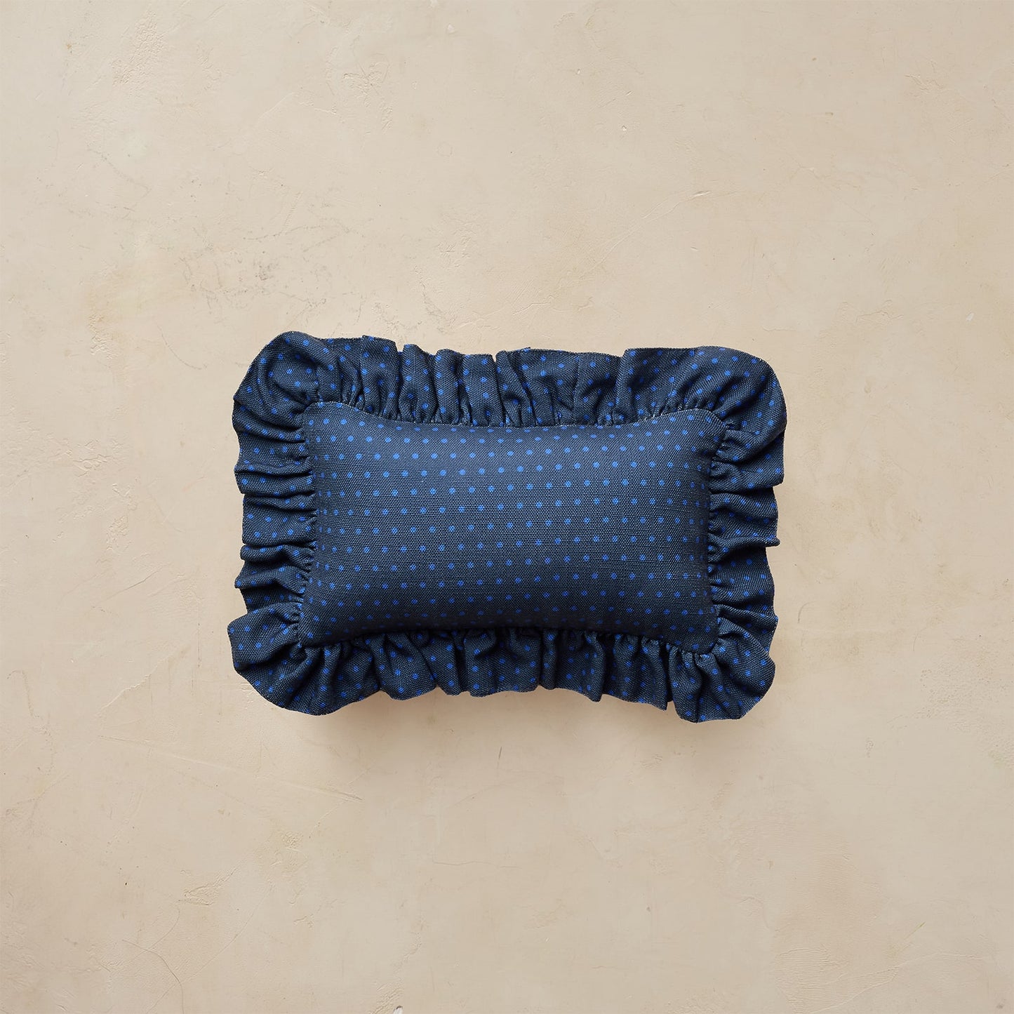 A rectangular navy blue pillow with a subtle polka dot pattern and ruffled edges, photographed from directly above on a textured beige background.