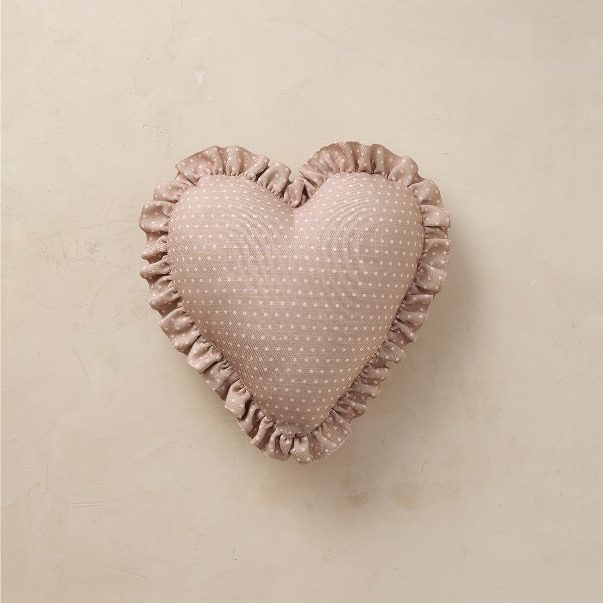 A heart-shaped decorative pillow in a dusty rose pink fabric with a small white polka-dot pattern and a ruffled satin border, displayed on a neutral beige textured background.