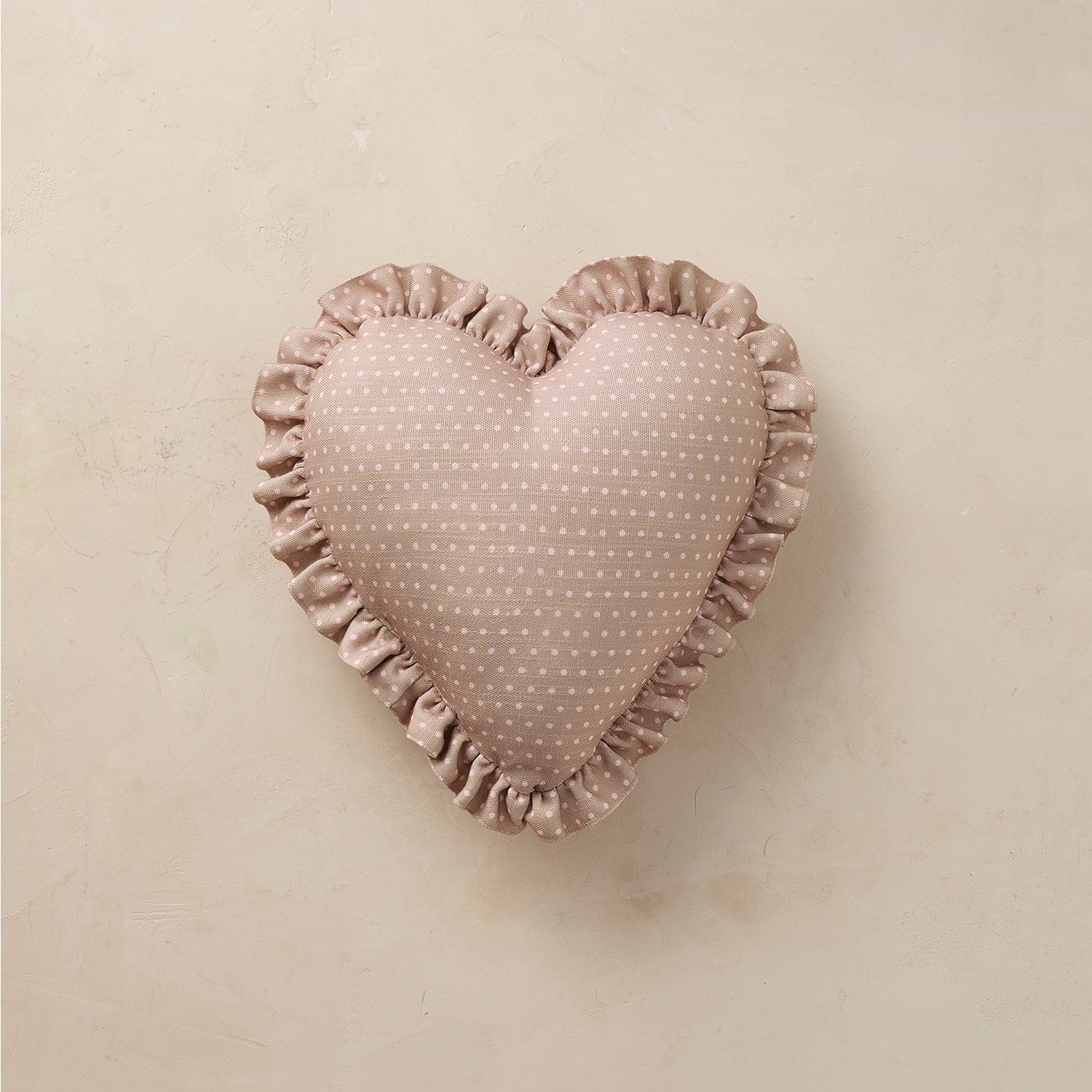 A heart-shaped decorative pillow in a dusty rose pink fabric with a small white polka-dot pattern and a ruffled satin border, displayed on a neutral beige textured background.