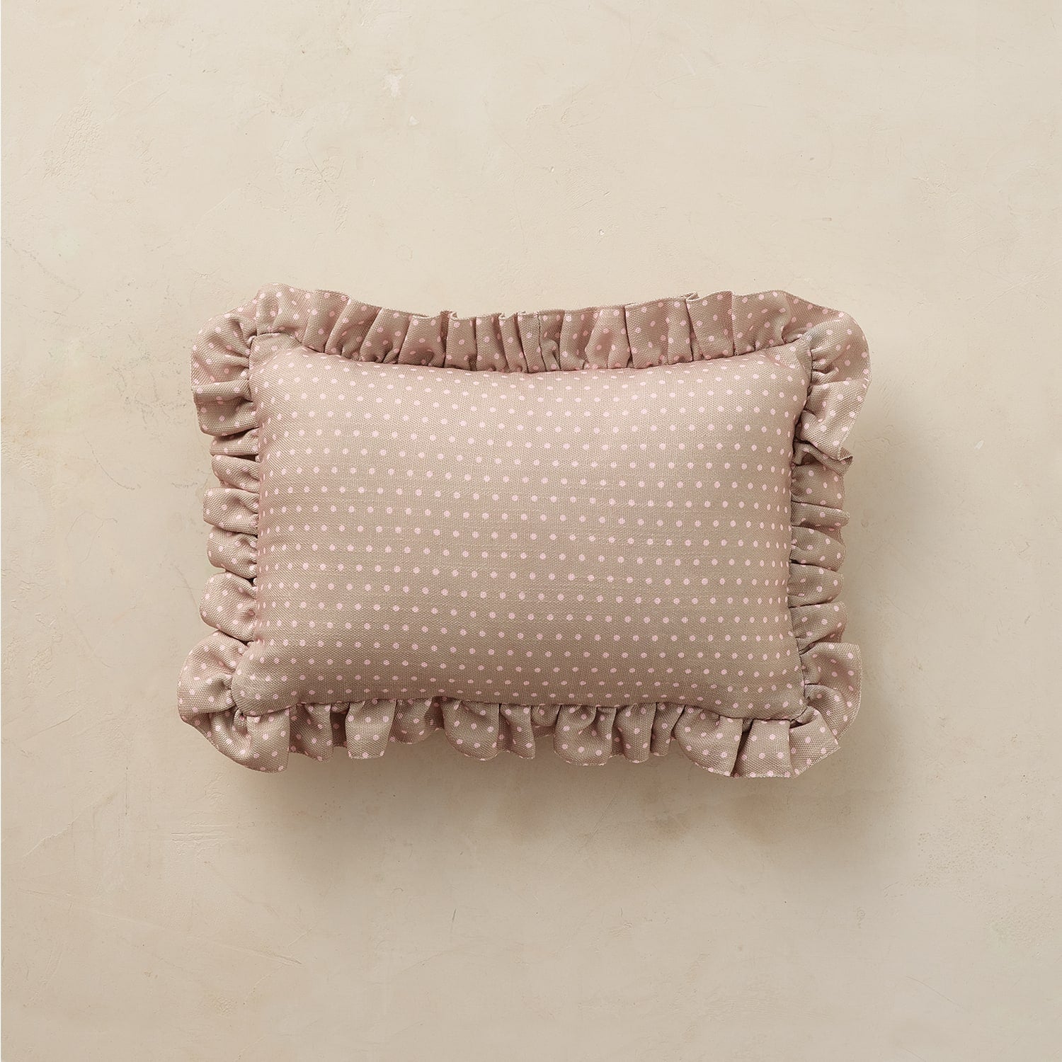 A rectangular throw pillow in a dusty rose velvet fabric with a white polka-dot pattern and ruffled trim, displayed on a neutral beige textured background.