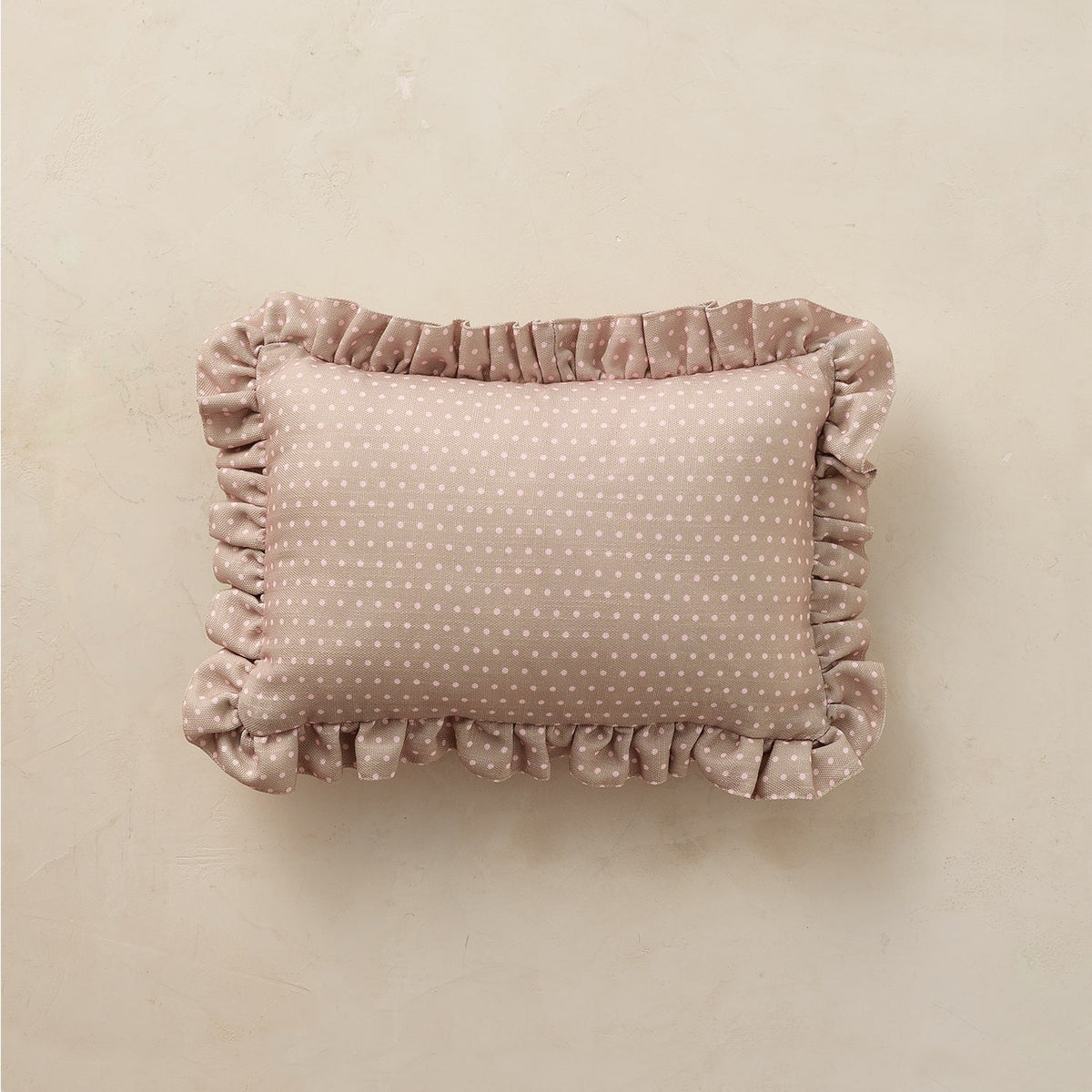 A rectangular throw pillow in a dusty rose velvet fabric with a white polka-dot pattern and ruffled trim, displayed on a neutral beige textured background.