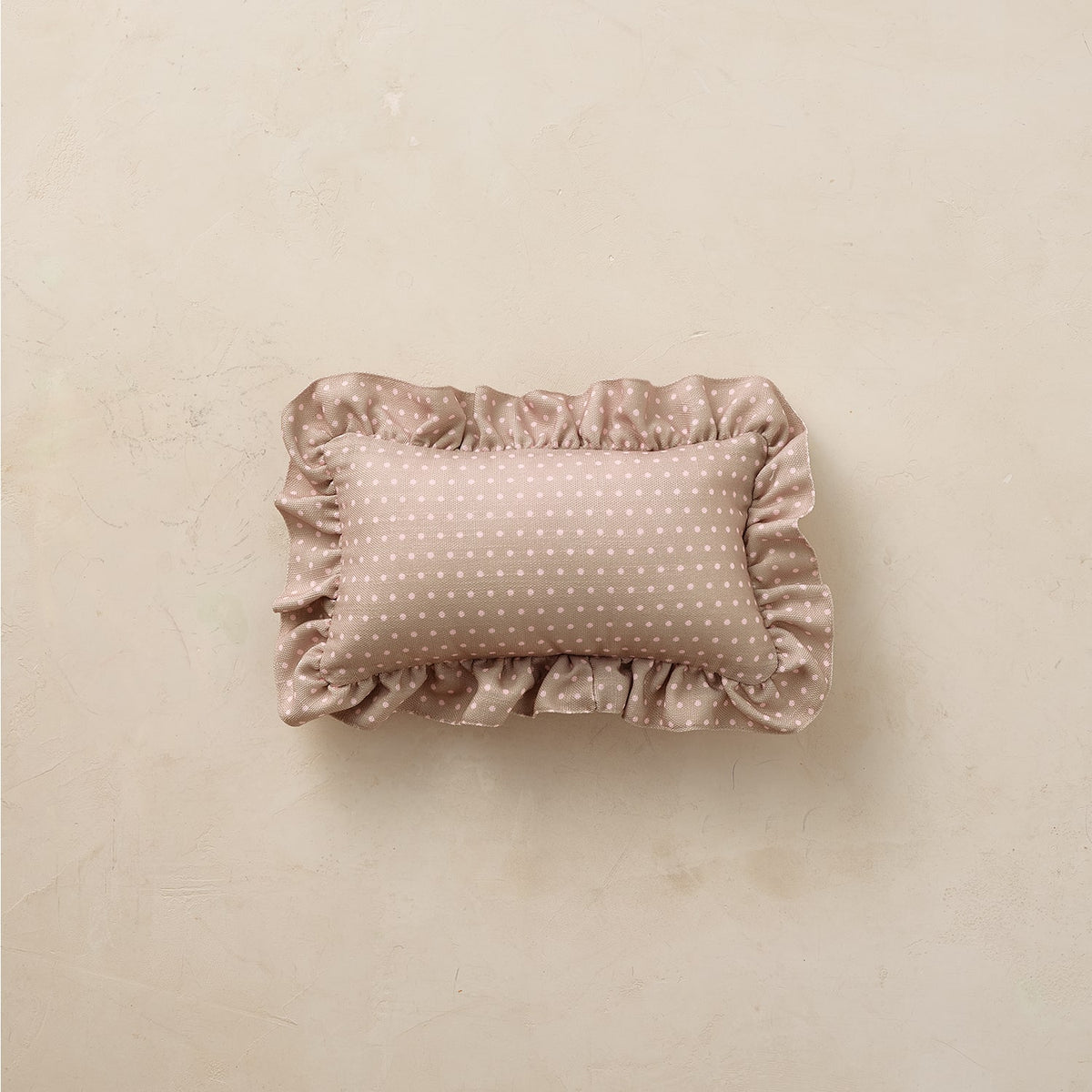 A small rectangular ruffled pillow in a dusty pink fabric with white polka dots, centered on a textured beige background.