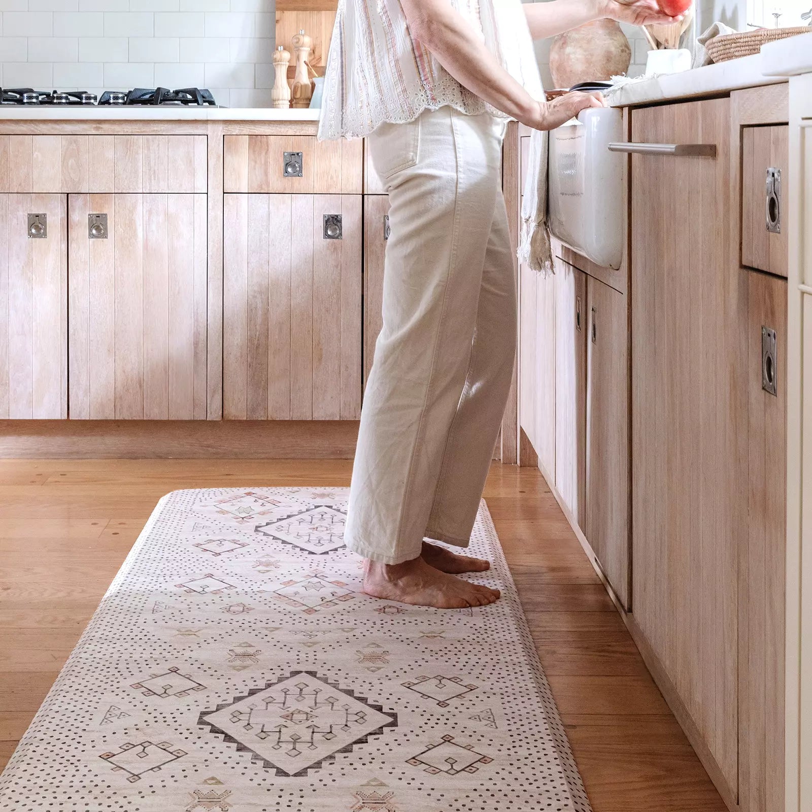 Nama Standing Mat in Ula Oat neutral boho print. Anti-fatigue kitchen mat shown in kitchen with woman washing dishes at the sink.