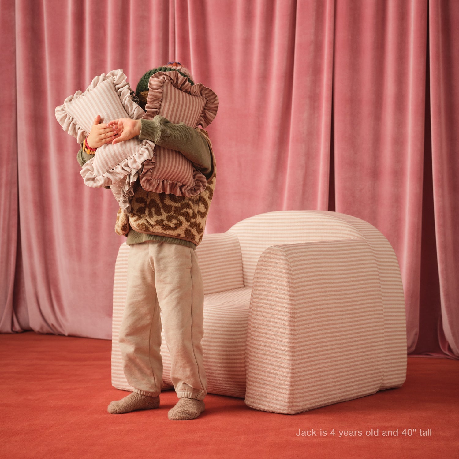 Child holding pillows in front of a pink curtain with a striped chair.