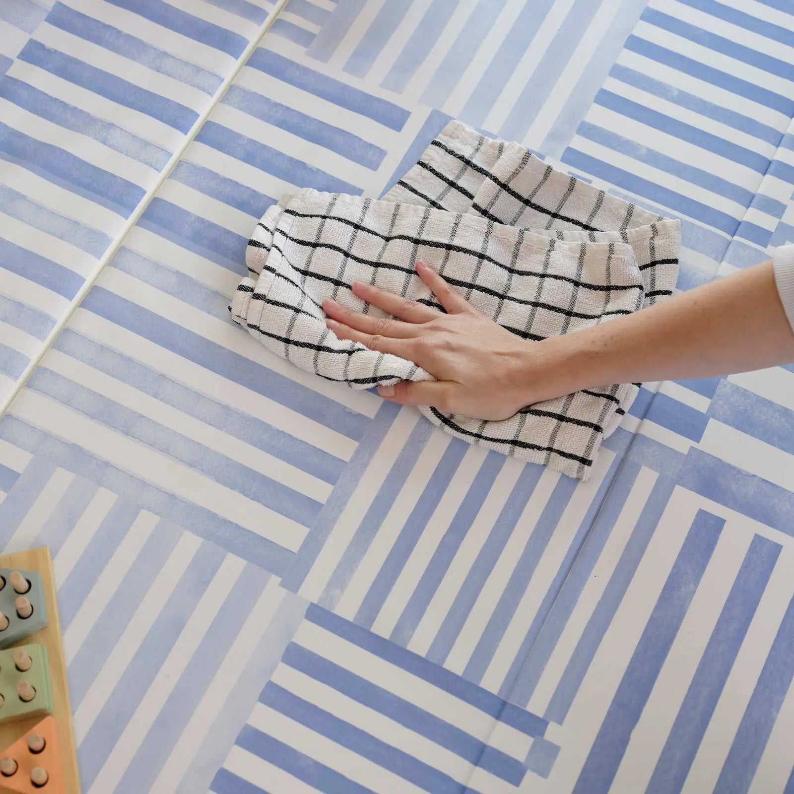 french blue inverted stripe blue and white tumbling mat. Shown with a hand wiping up a spill with a towel and a wooden toy peaking out of the lower left hand corner