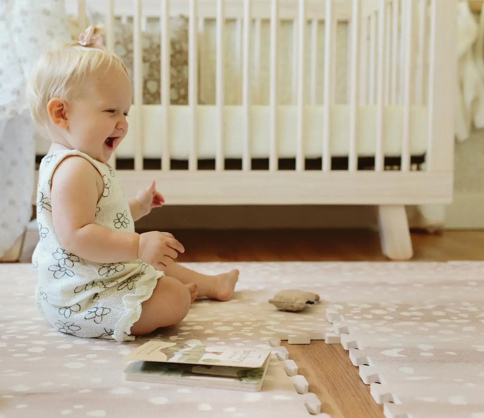 Fawn brown neutral animal print play mat with 1 tile exposd shown in nursery with laughing baby sitting on the mat with a book and toy