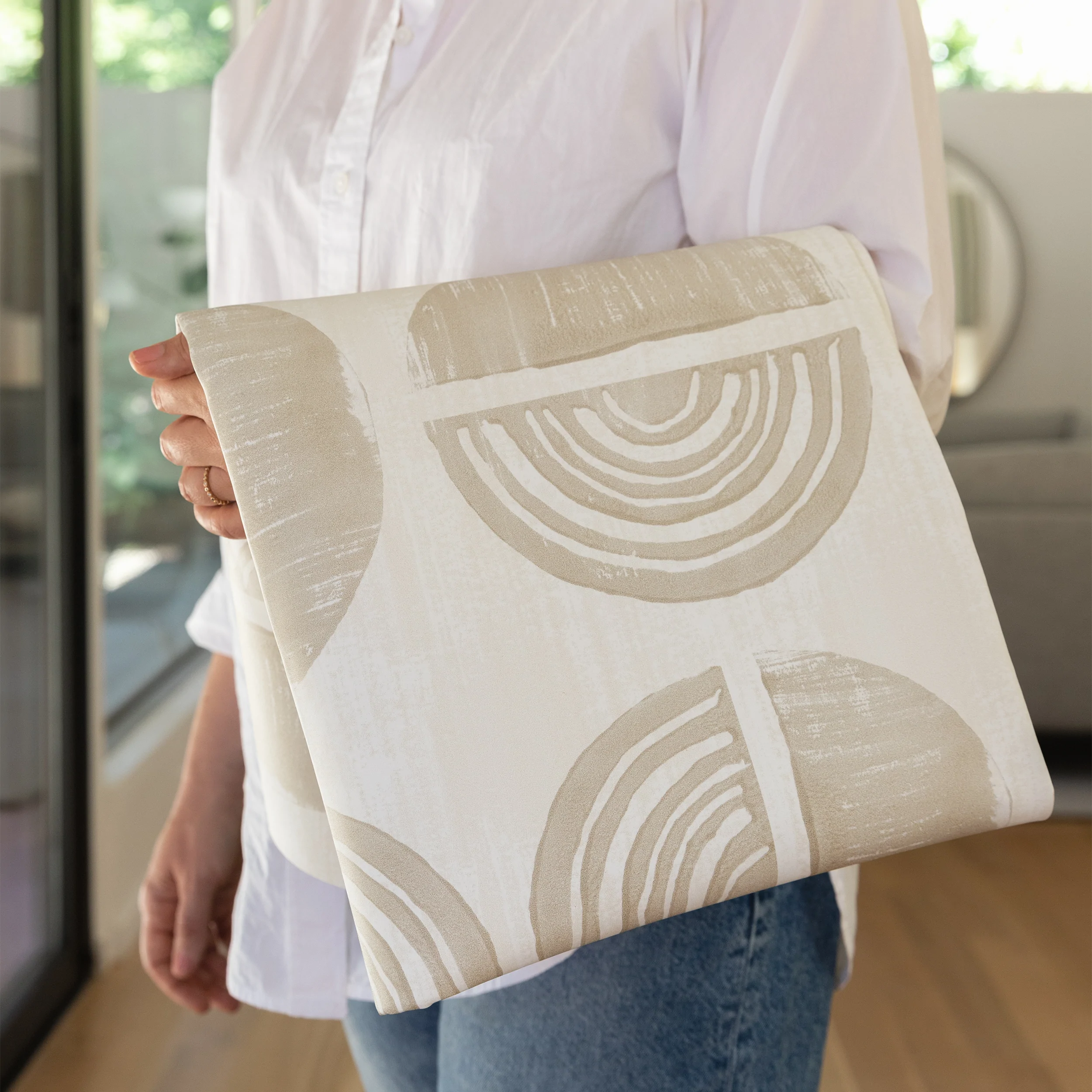 Ada modern minimalist baby high chair mat in Pebble taupe and off white shown folded up being carried by a woman.
