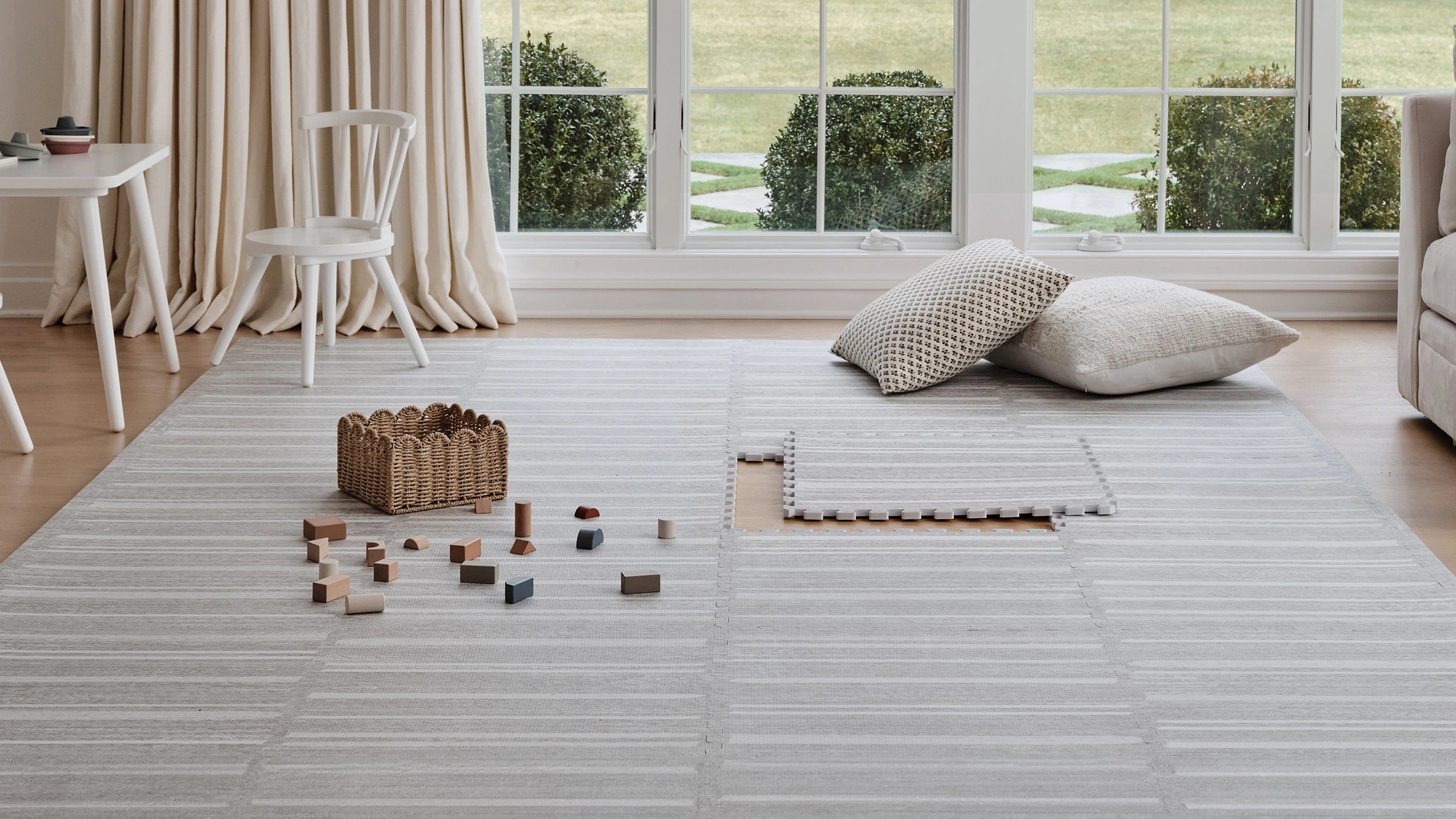 Rowan birch beige and white stripe play mat shown in a living room with basket of blocks and pillows on top of the mat. One tile exposed in the middle