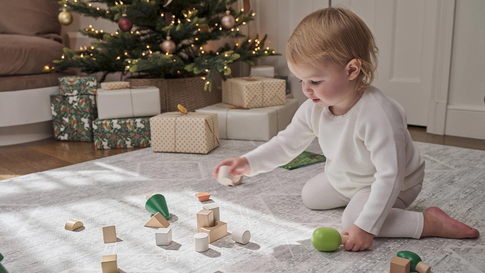 Ula gray boho play mat shown in a living room decorated with a christmas tree and wrapped presents with toddler girl playing with wooden blocks on the mat