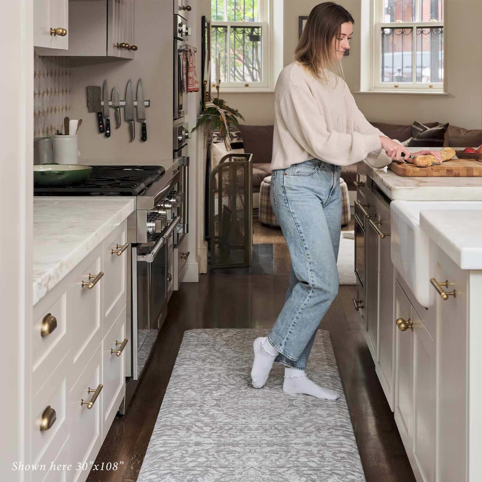 Emile earl grey gray and cream floral standing mat shown in kitchen in size 30x108 with woman standing on the mat cutting bread on a cutting board on the counter