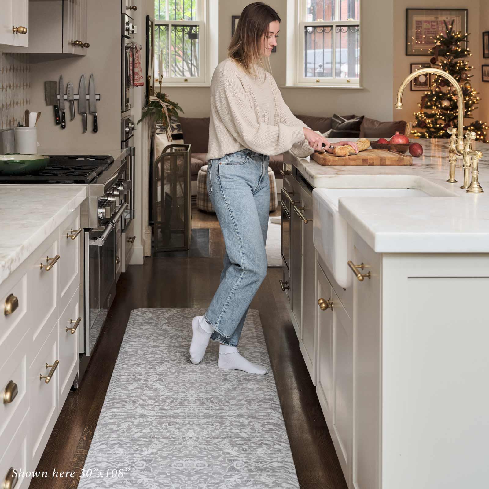 Emile earl grey gray and cream floral standing mat shown in kitchen in size 30x108 with woman standing on the mat cutting bread on a cutting board on the counter