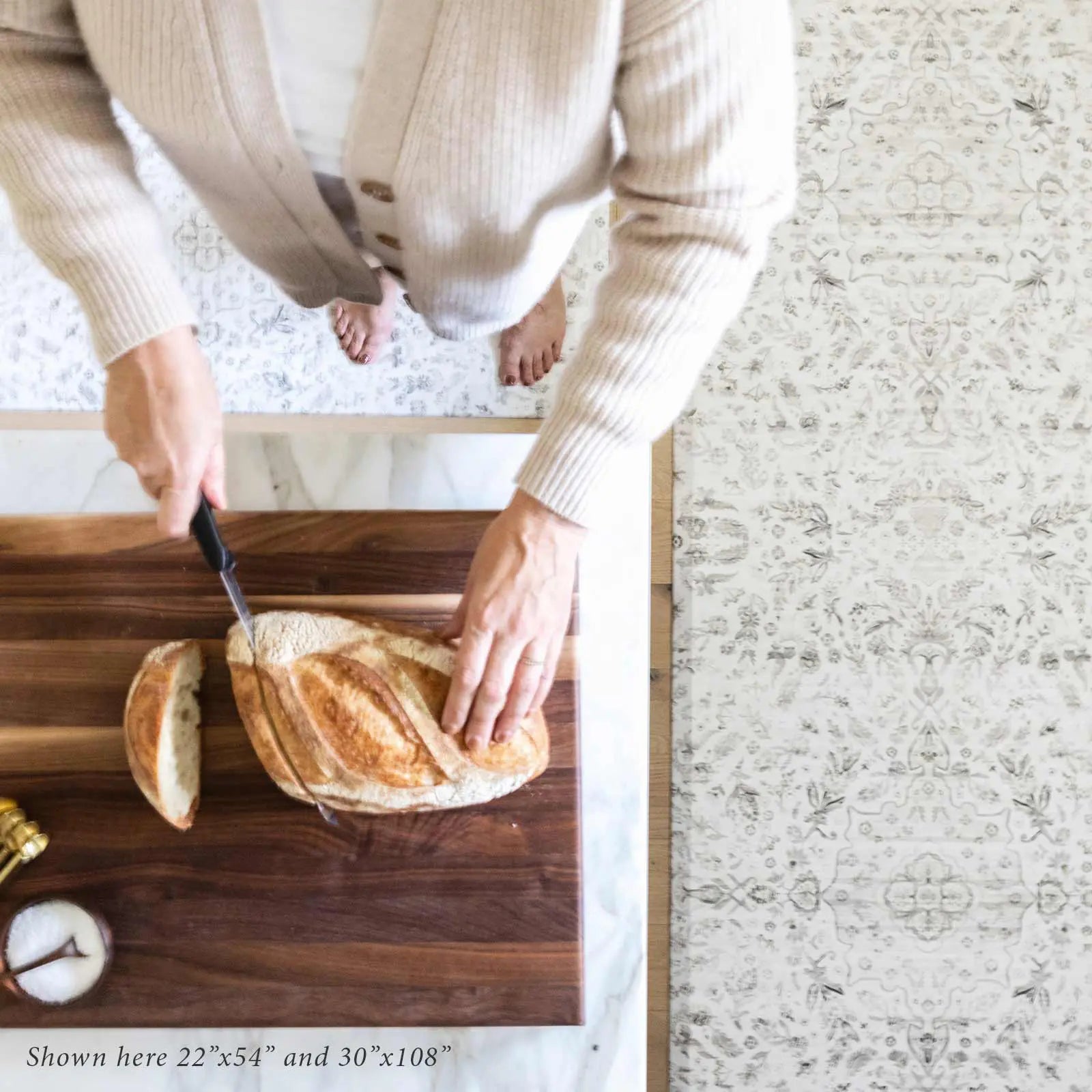 Emile latte beige and gray floral boho kitchen mat shown in sizes 22x54 and 30x108 with woman cutting a loaf of bread at kitchen counter