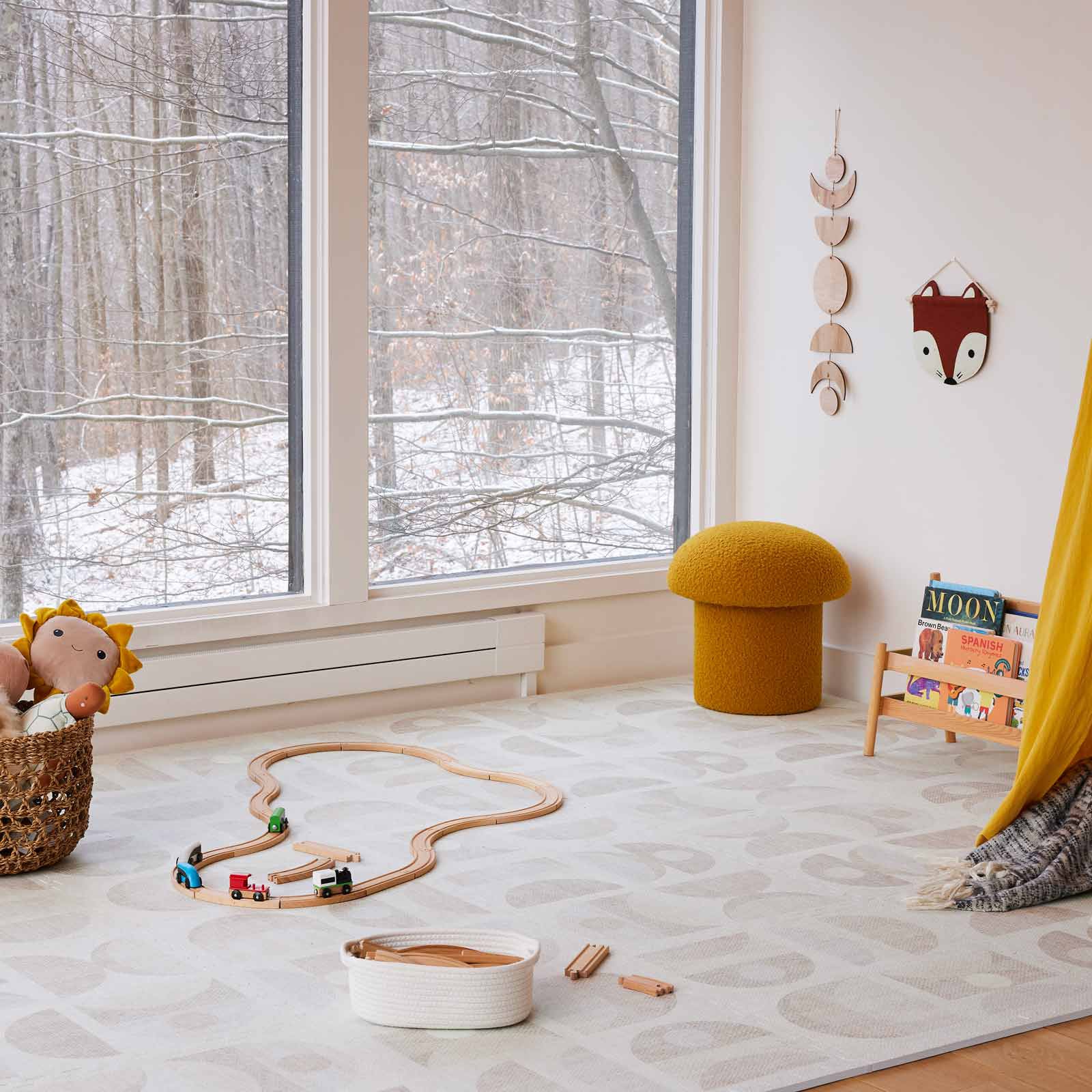 Luna sandstone neutral geometric print play mat shown in play room with wooden train track toys, mustard mushroom ottoman in the corner, wooden train tracks laid out on the mat, a basket of stuffed animals, and a small wooden book stand