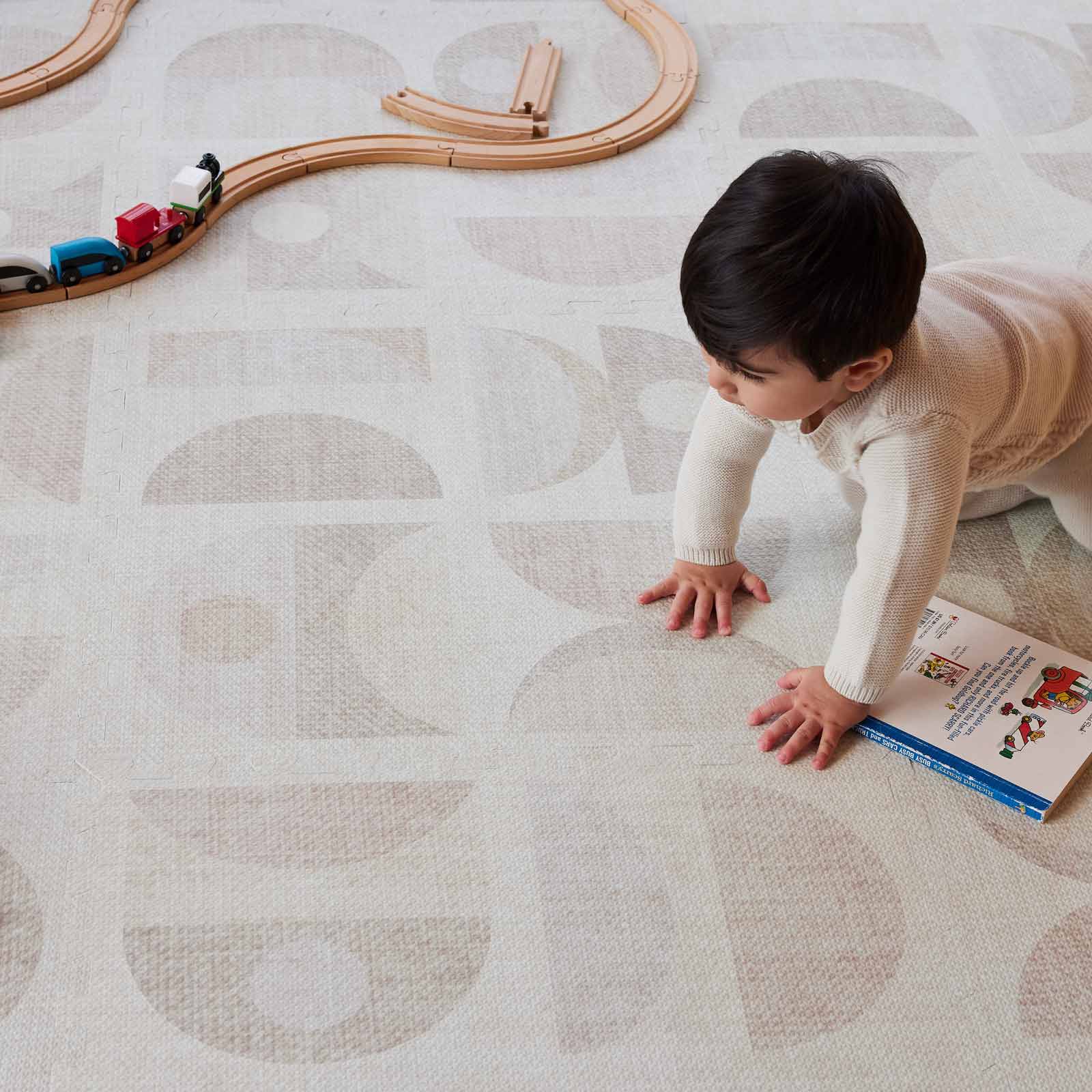Luna sandstone neutral geographic print play mat with baby boy crawling over a book on the mat with a wooden train track in the corner of the mat