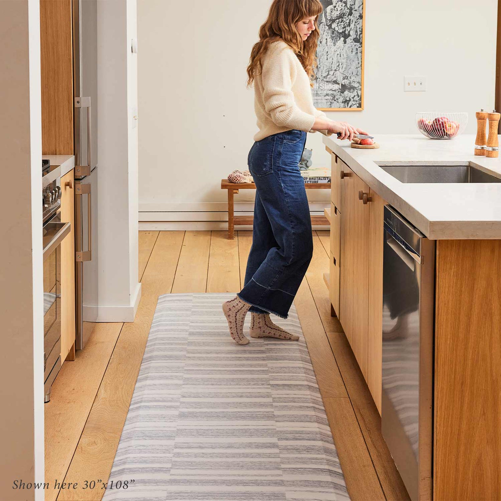 Sutton stripe heather gray and white inverted stripe standing mat shown in light wood tone kitchen with woman standing on size 30x108 cutting up fruit at the counter