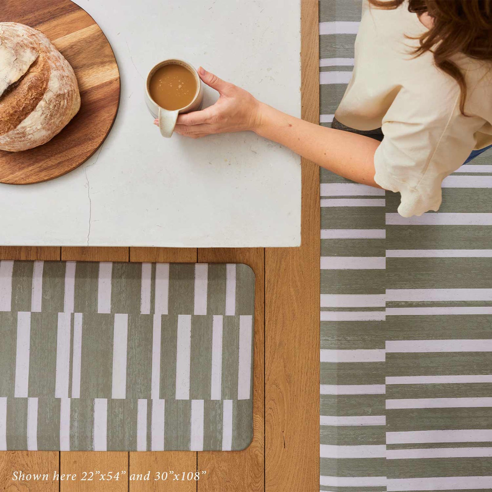 sutton stripe basil green and white inverted stripe standing mat shown from in sizes 22x54 and 30x108 with woman standing on the mat at kitchen counter holding a cup of coffee next to a loaf of bread