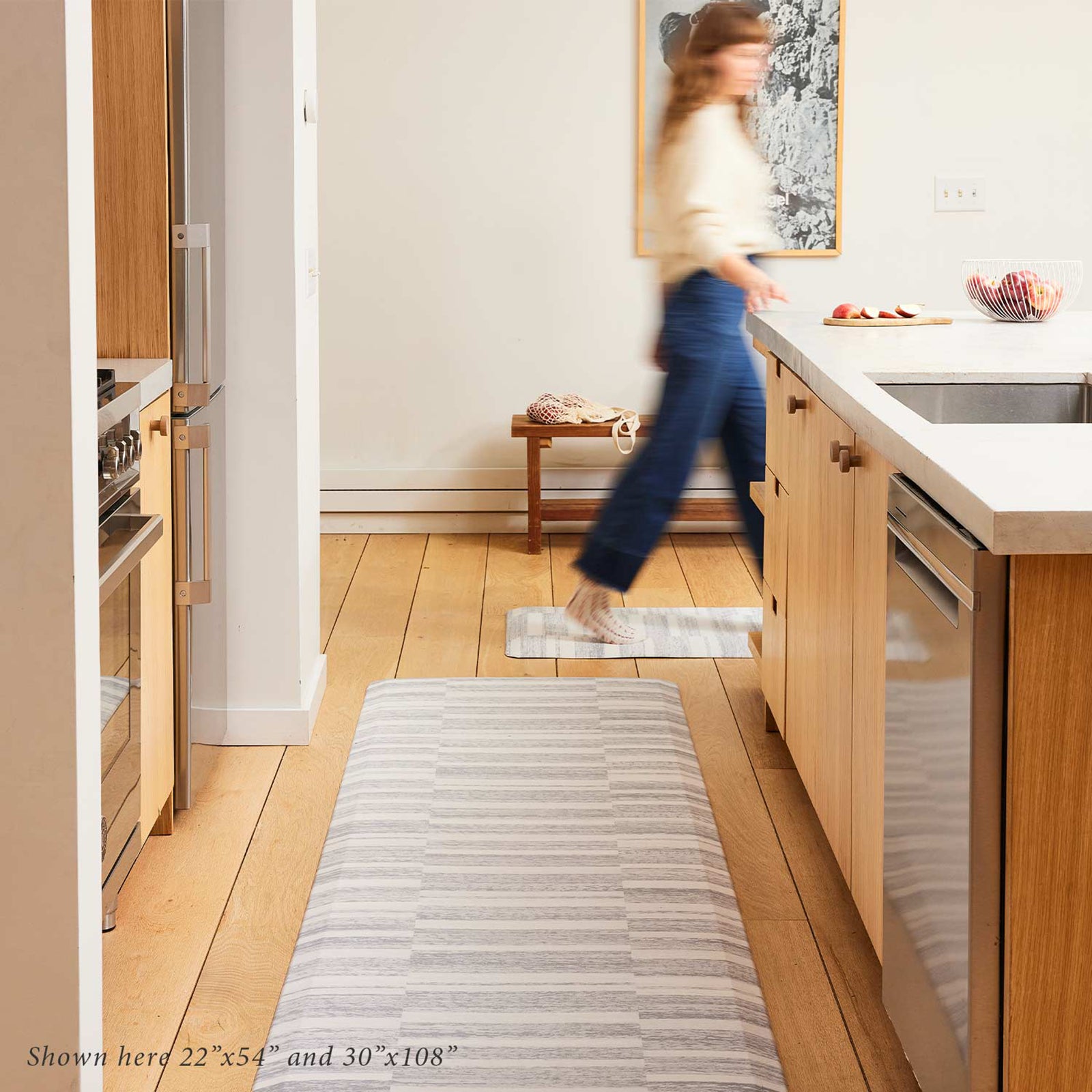 Sutton stripe heather gray and white inverted stripe standing mat shown in light wood tone kitchen in sizes 22x54 and 30x108 with woman walking past in the background