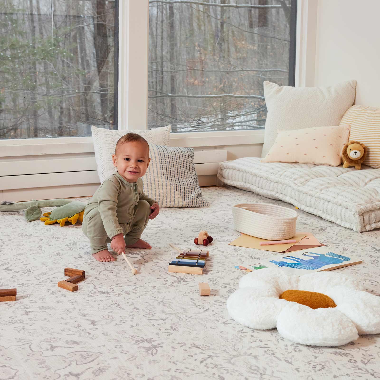 Emile latte neutral floral play mat shown in play room with baby boy playing with a xylophone with plush toys and pillows on top of the mat
