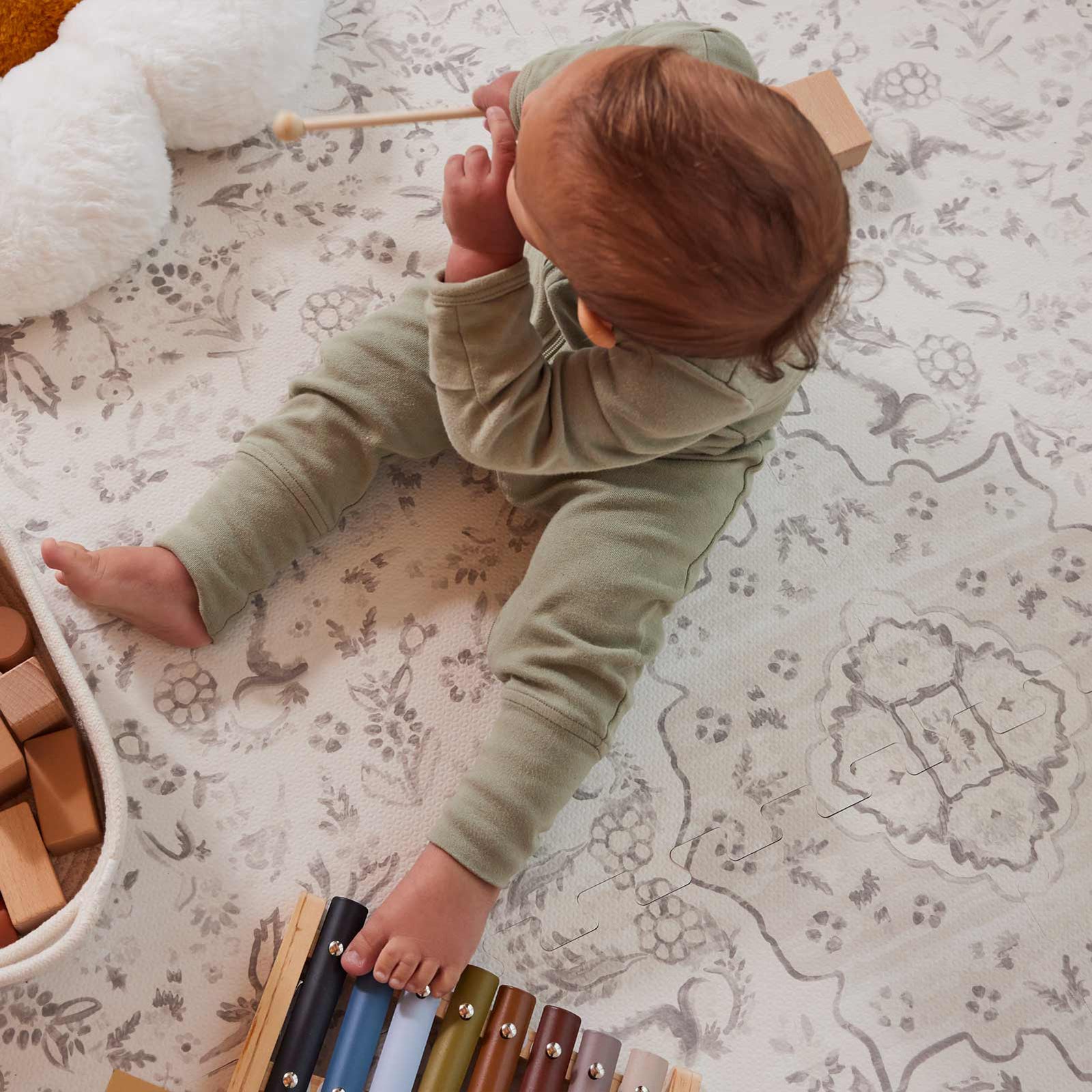 Emile latte neutral floral play mat shown from above with baby boy sitting in front of a plush toy, basket of wooden blocks, and a toy xylophone