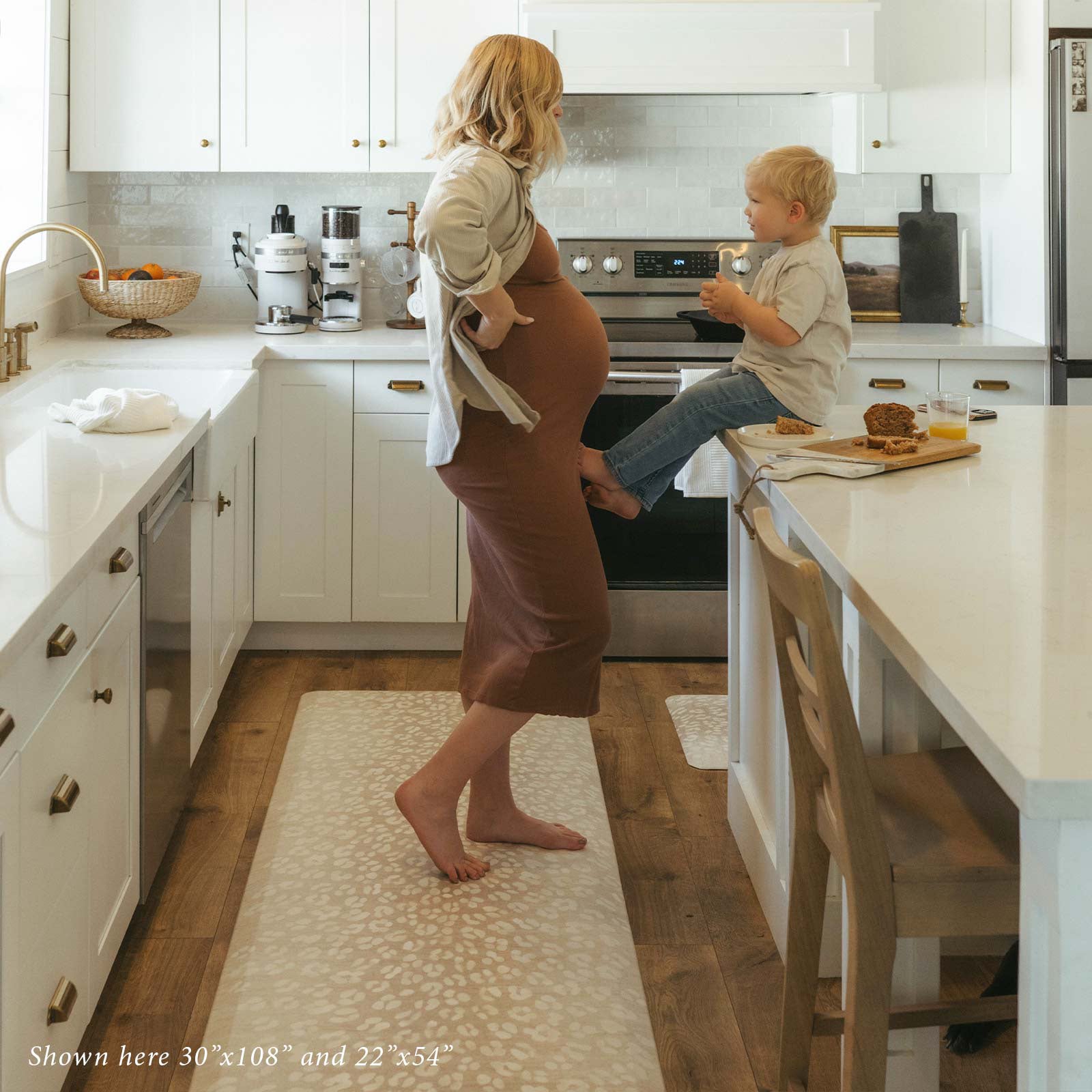 Della Salt neutral abstract animal print standing mat, showing size 30x108 in a kitchen with pregnant mom standing on the mat in front of toddler boy sitting on the counter eating a snack