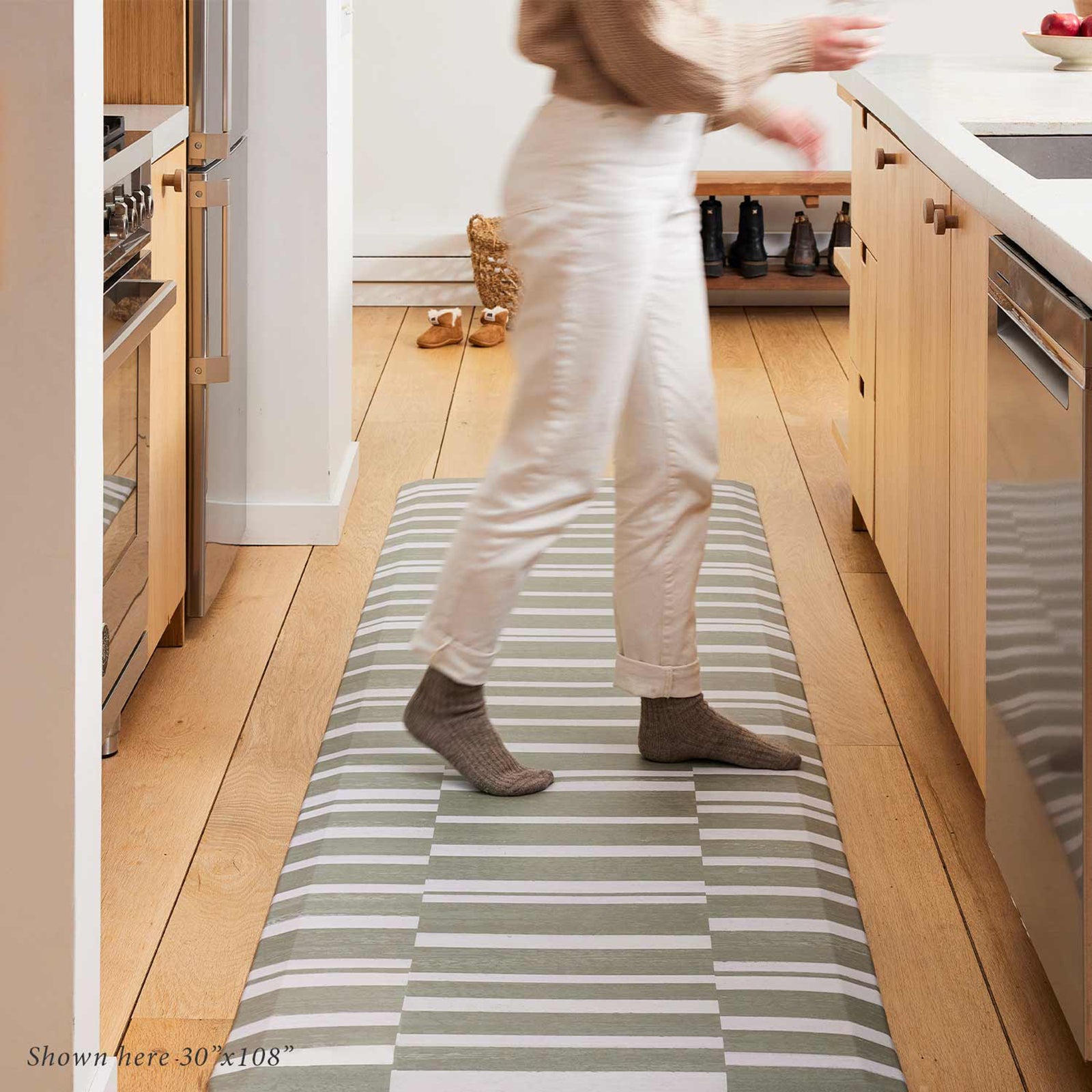 sutton stripe basil olive green and white inverted stripe standing mat shown in kitchen with woman wearing white jeans and gray socks standing on the mat moving toward the sink