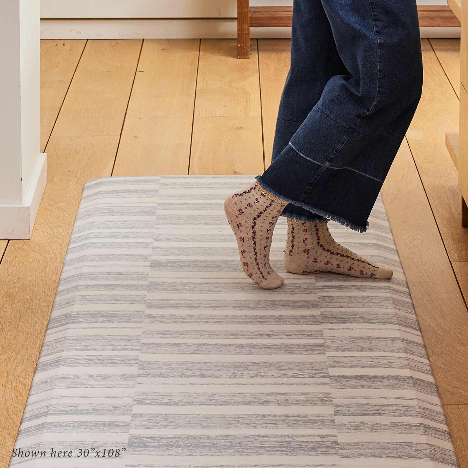Sutton stripe heather gray and white inverted stripe standing mat shown in size 30x108 cropped to show the feet of a woman standing on the mat wearing floral socks and blue jeans