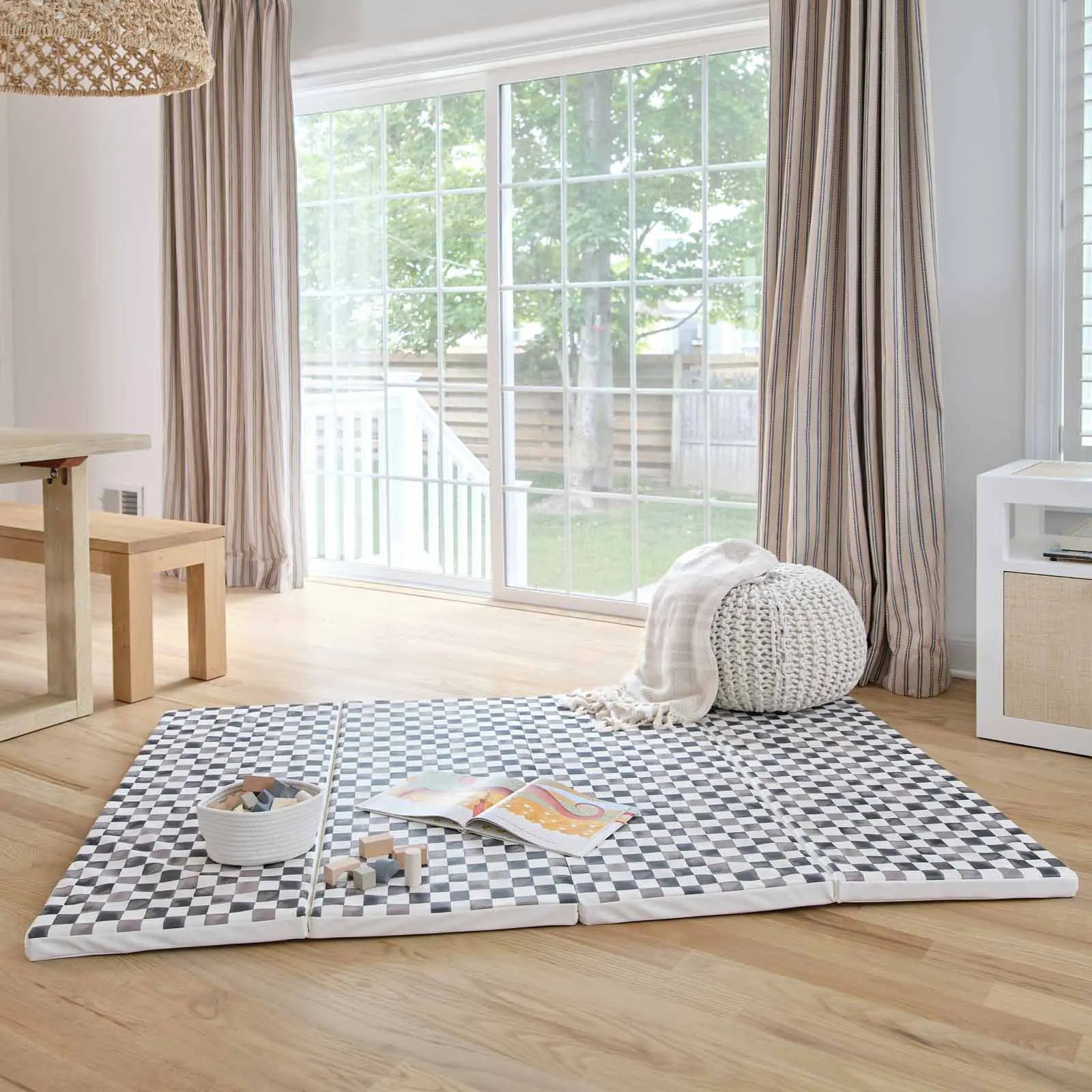 Tuxedo black and white checker print tumbling mat shown in open living space with a pouf, blanket, basket of blocks, and a book on the mat