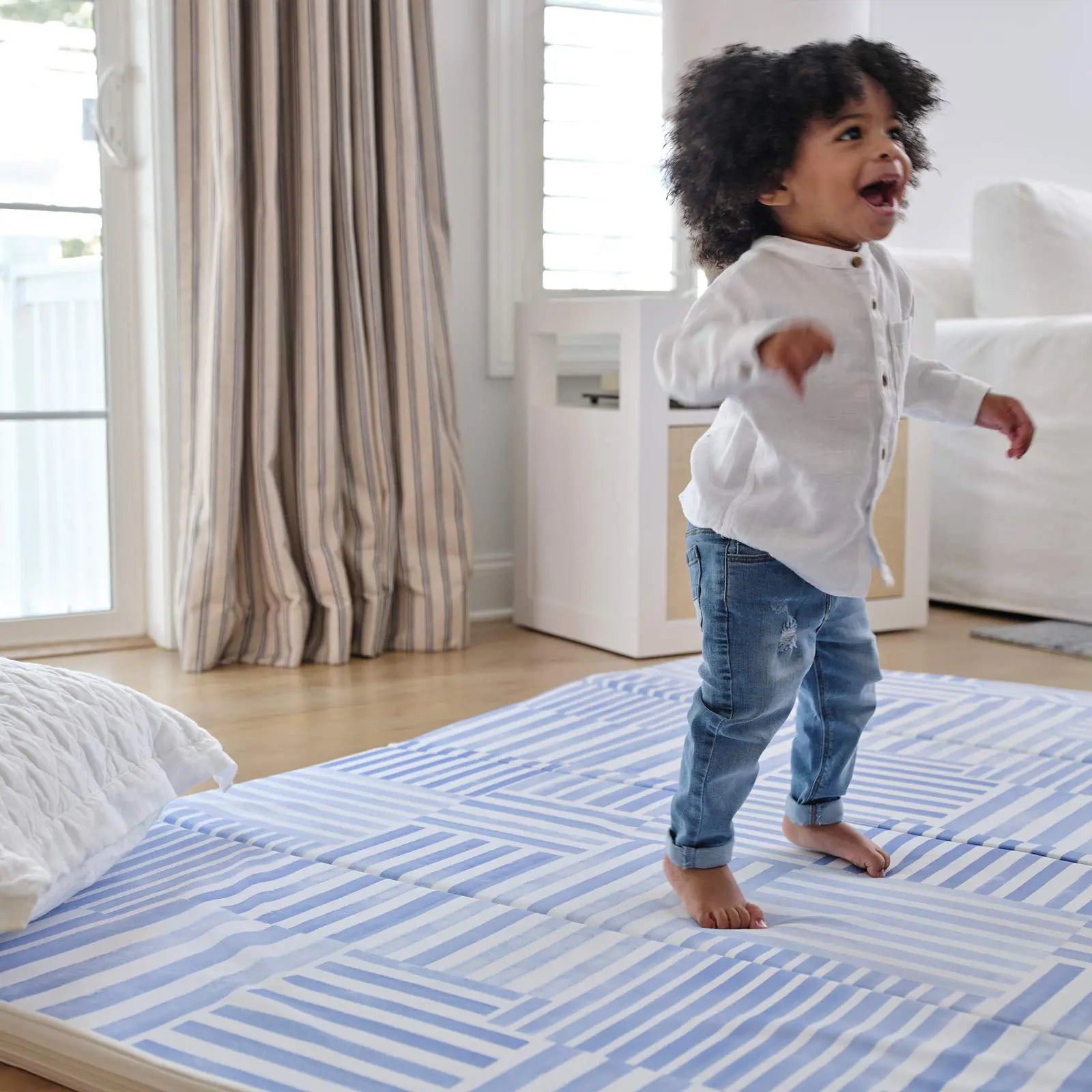 French blue and white inverted stripe tumbling mat shown with toddler smiling and jumping on the mat with a pillow on the corner of the mat