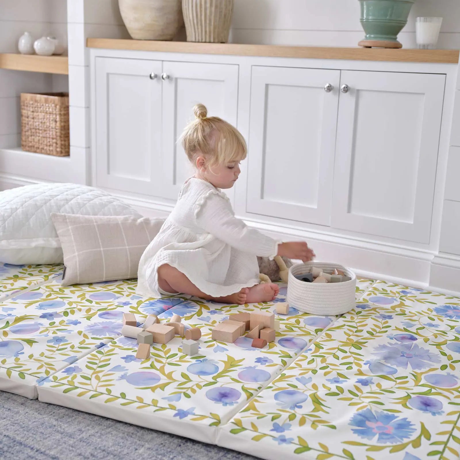 Bluebell blue green and white floral tumbling mat shown in a living room with toddler girl sitting on the mat with pillows behind her playing with wooden blocks