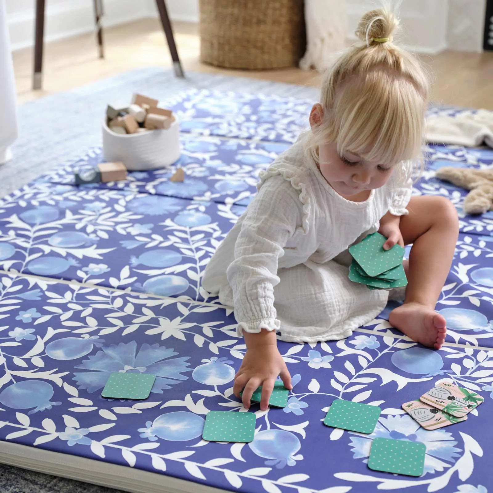 Deep sea blue and white floral tumbling mat with toddler girl sitting on the mat playing a memory card game with basket of blocks and plush toys on the mat in the background