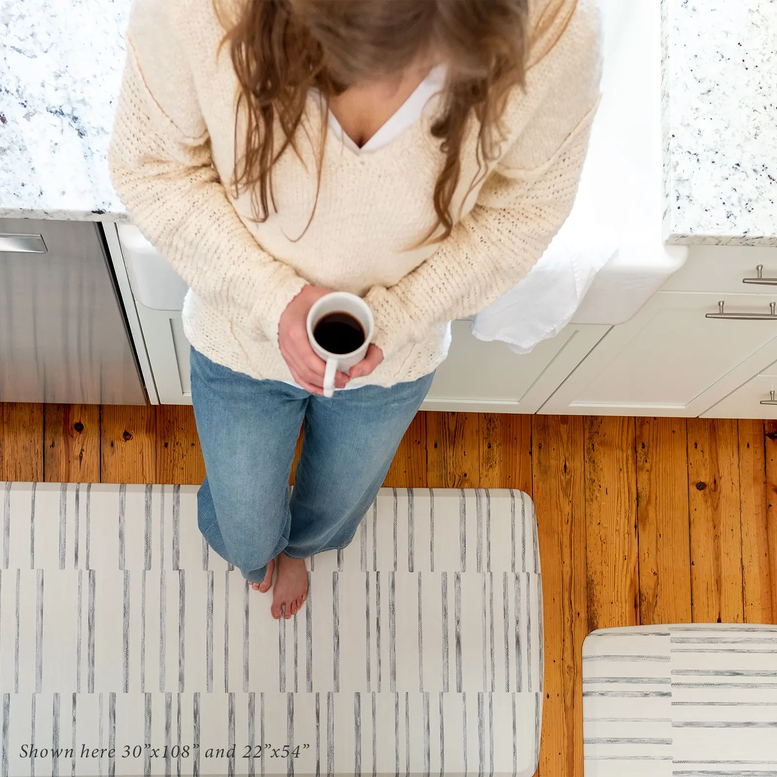Gray and white inverted stripe kitchen mat shown from above in kitchen with woman drinking a cup of coffee standing on size 30x108