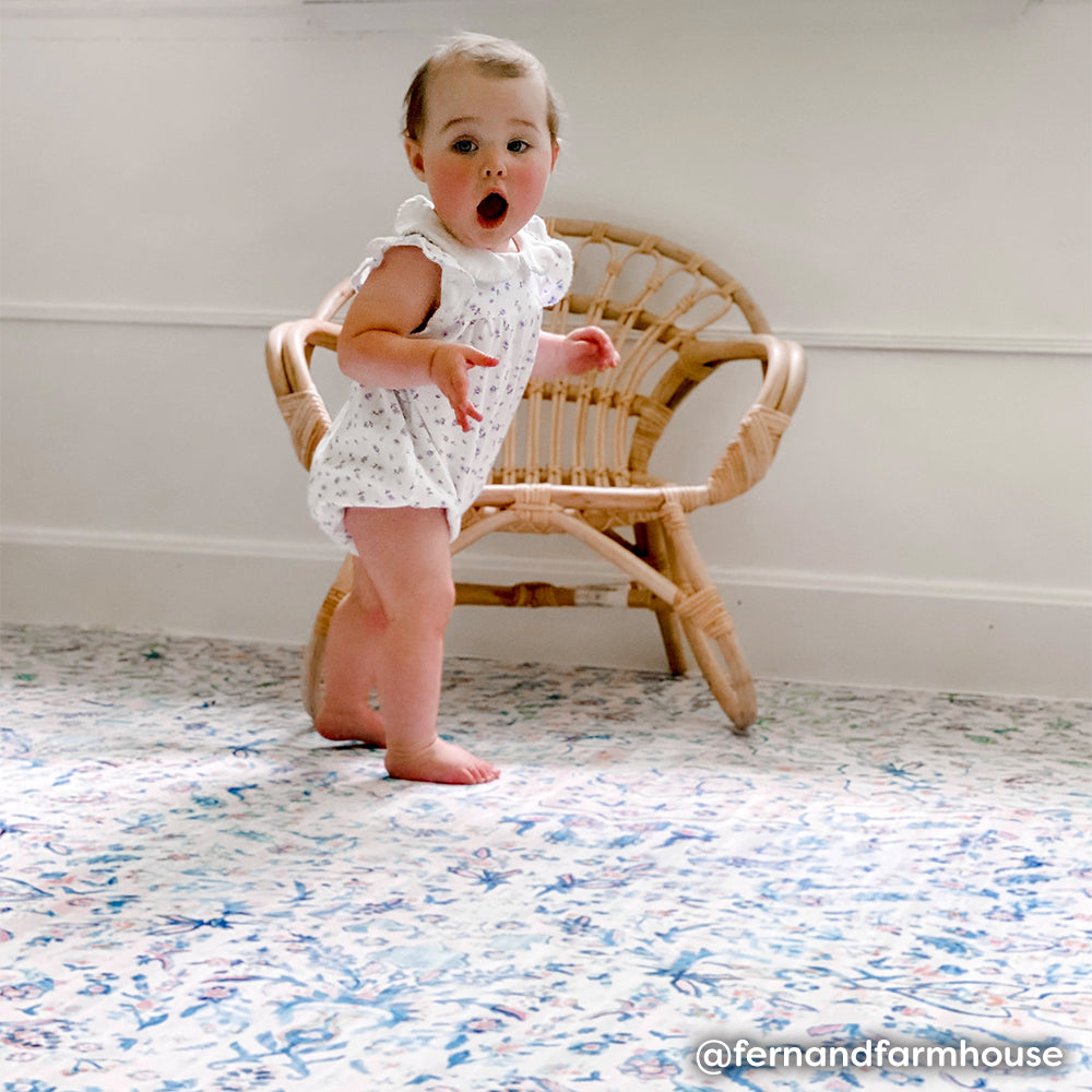 Blue and blush vintage floral print baby play mat shown with baby standing next to chair making a surprised face.
