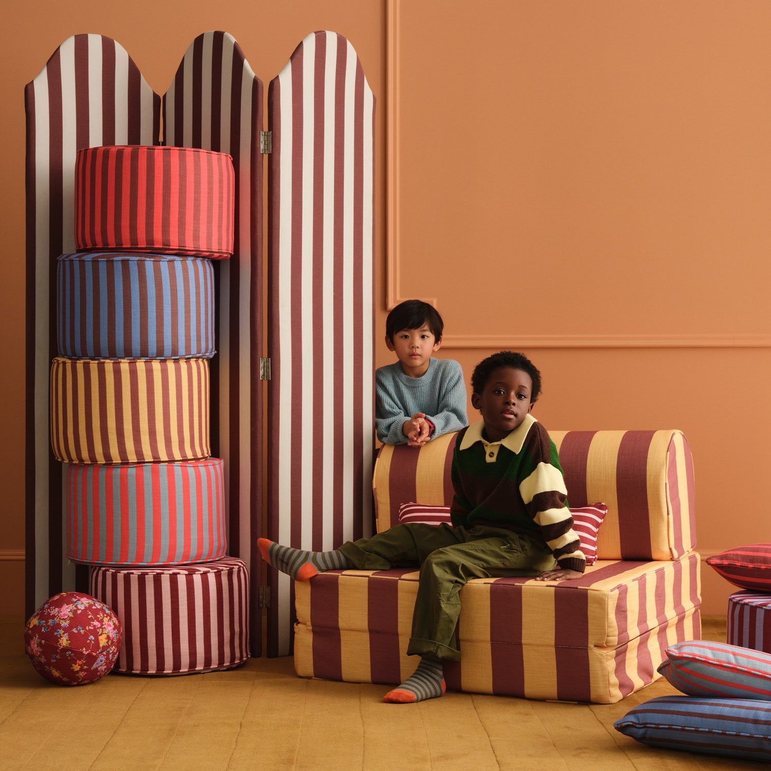 Two children sitting on a striped chair with colorful cylindrical objects in a room.
