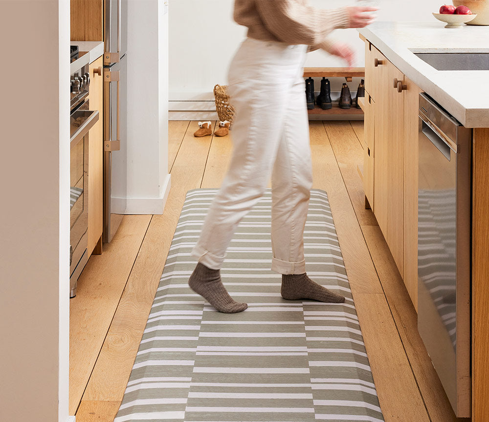 sutton stripe basil olive green and white inverted stripe standing mat shown in kitchen with woman wearing white jeans and gray socks standing on the mat moving toward the sink