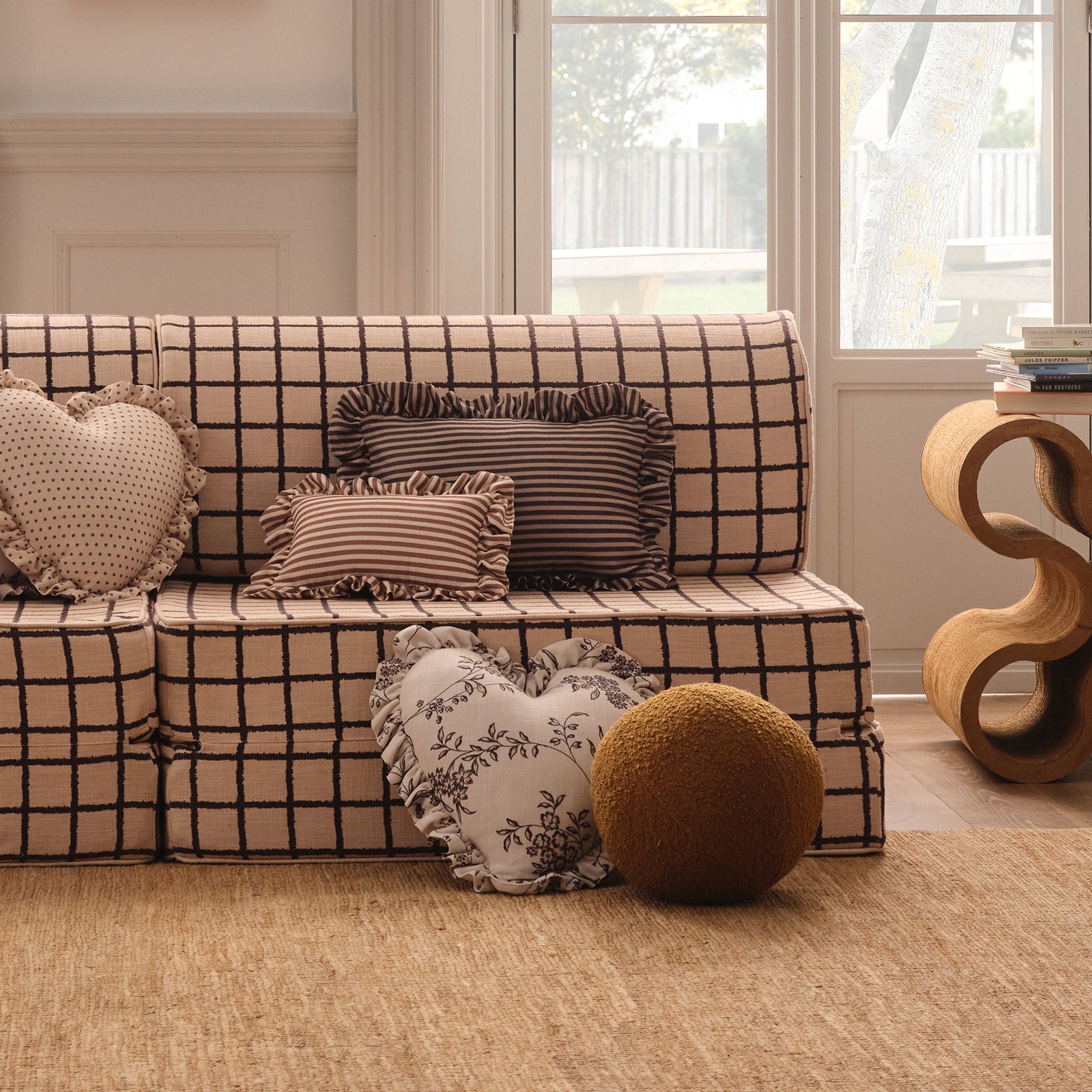 A beige and black checkered sofa in a warm, neutral-toned living room, styled with textured throw pillows, a heart-shaped floral cushion, and a round brown pouf on a woven rug.