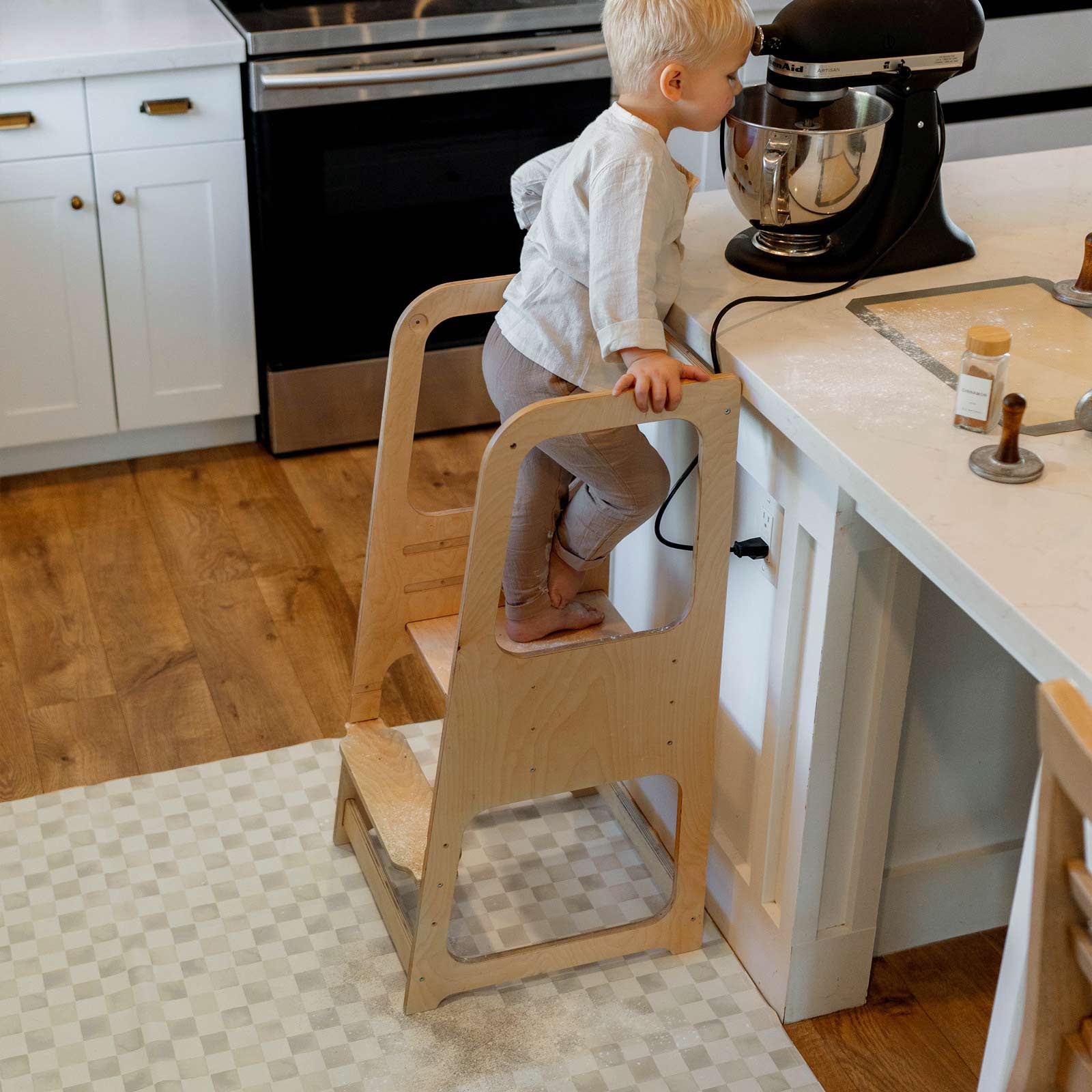 Checker almond tonal beige geometric print high chair mat shown with toddler standing on a stool making cookies at the kitchen counter