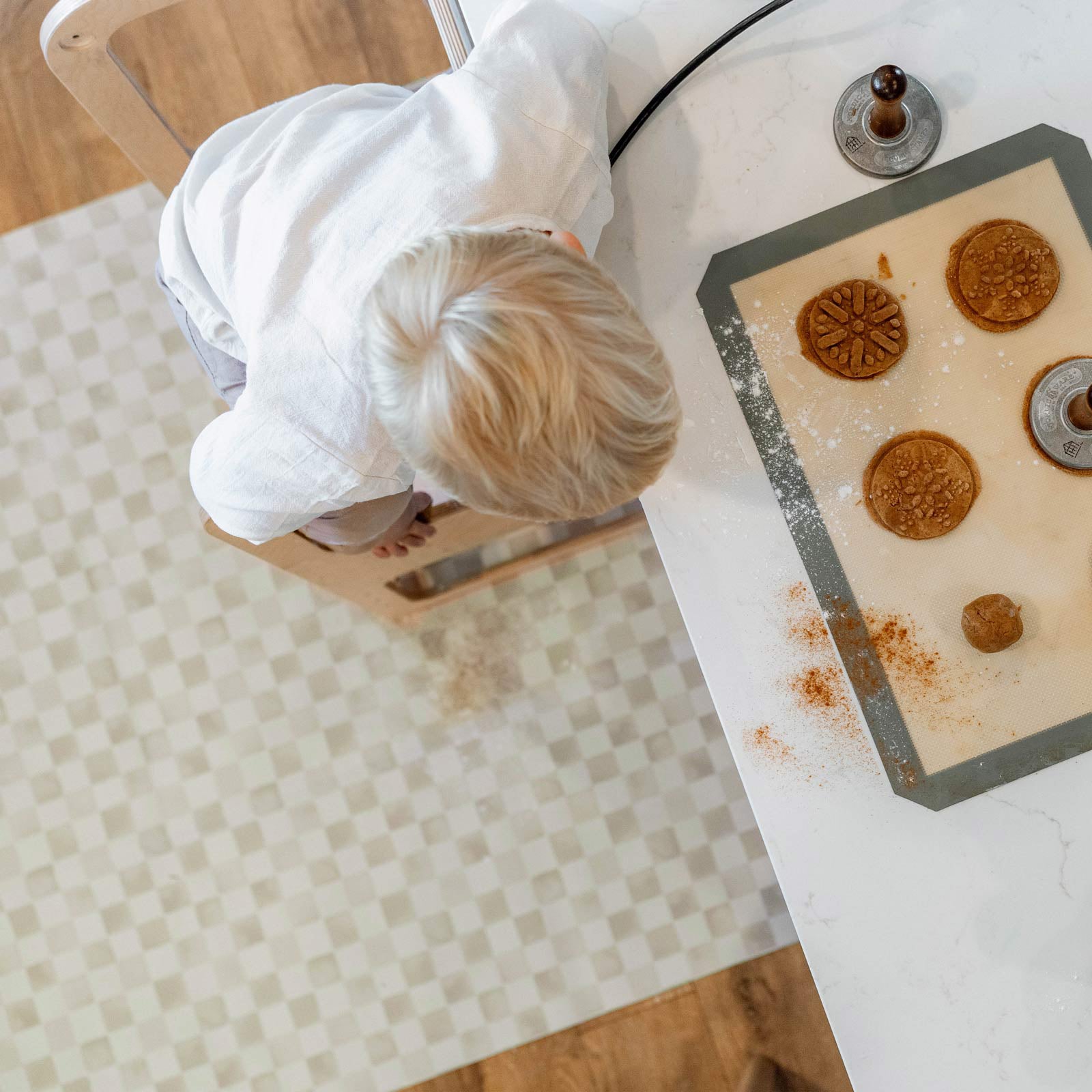 Checker almond tonal beige geometric print high chair mat shown from above in kitchen with toddler standing on a stool making cookies at the counter