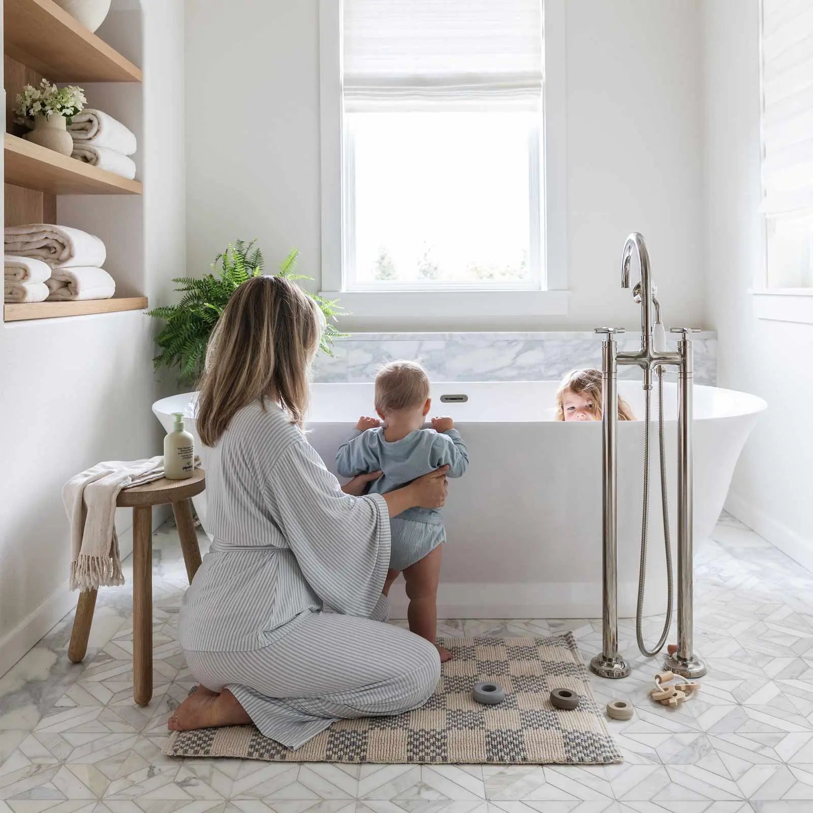 Harbor grey grey and white checker print bath mat shown with moms feet standing on the mat in front of a vanity and baby pulling itself up on the vanity