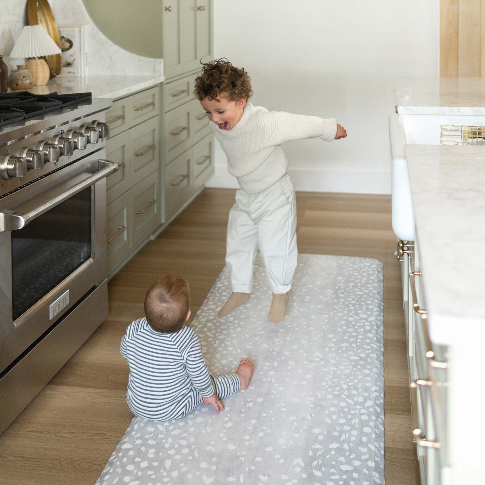 Fawn silver gray animal print standing mat shown in kitchen in size 30x108 with toddler boy jumping up and baby boy sitting on the mat looking at the older boy.