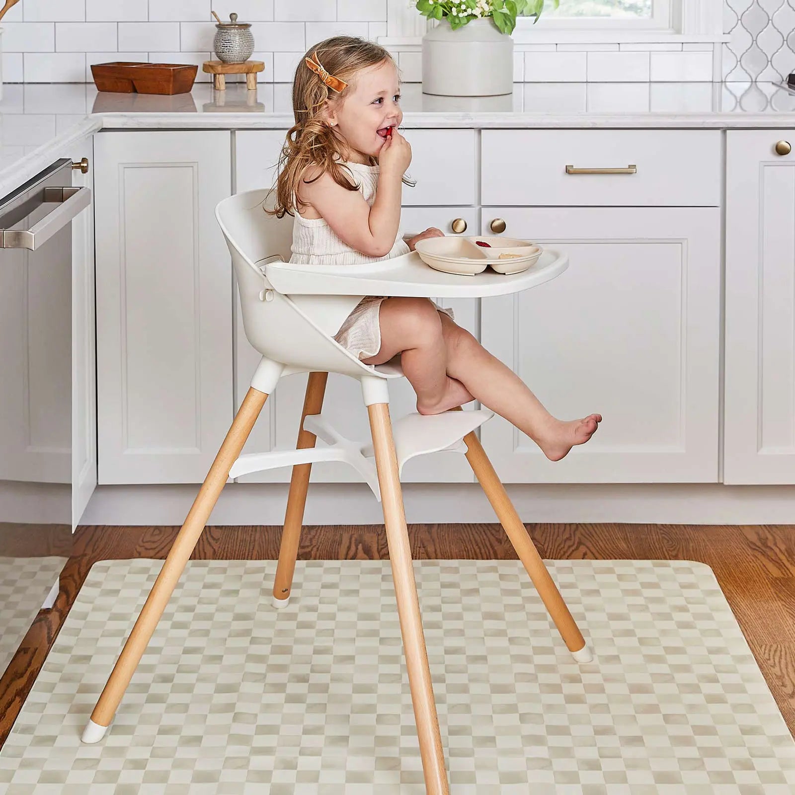 Checker almond beige geometric print high chair mat shown in a kitchen underneath a highchair with a toddler girl sitting in the chair smiling and eating fruit