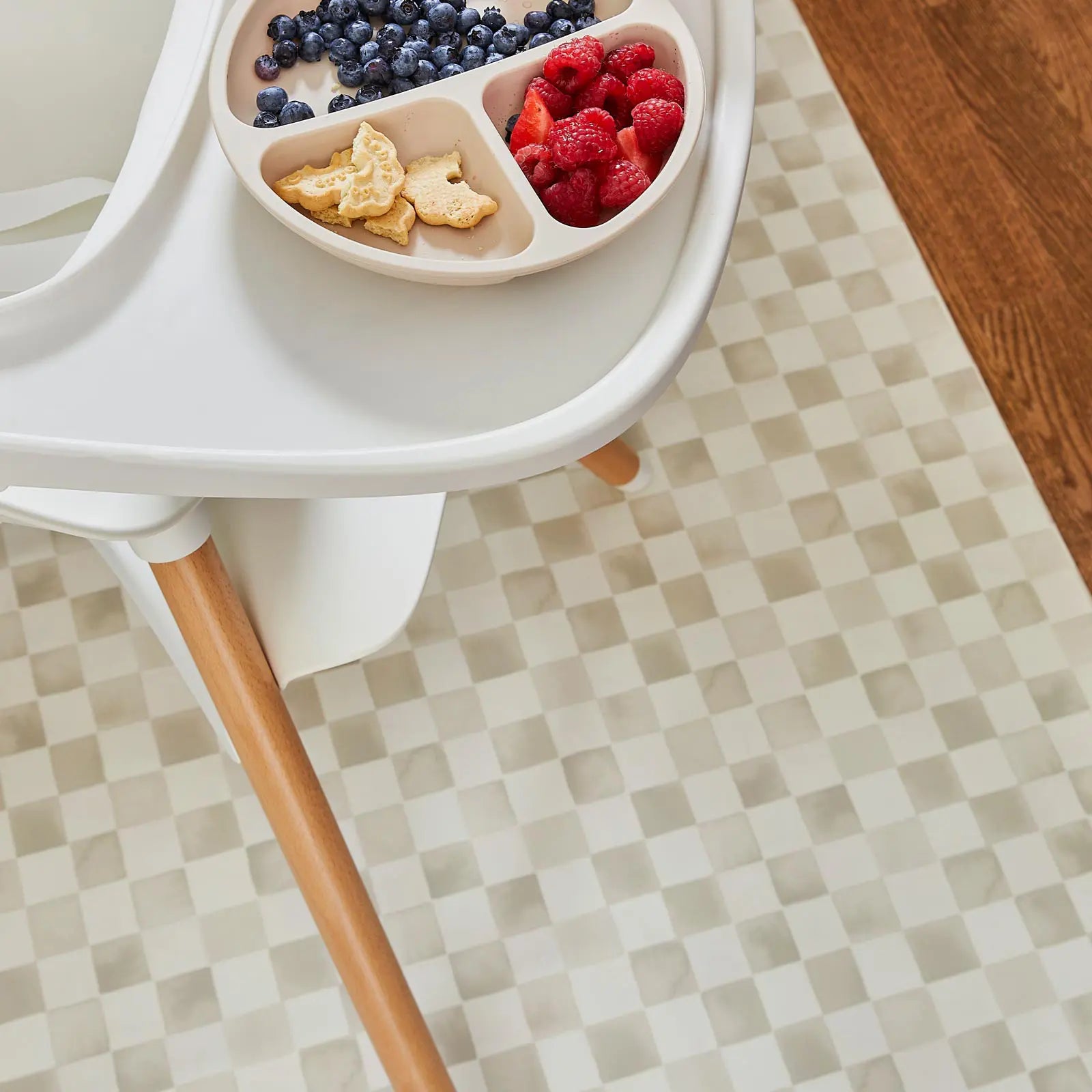 Checker almond beige geometric print high chair mat shown beneath a highchair with fruit and cookies on the tray