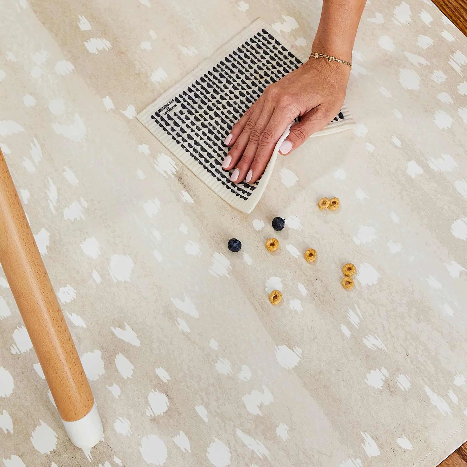 Fawn brown and white animal print high chair mat shown with womans had wiping up spilled blueberries and cereal